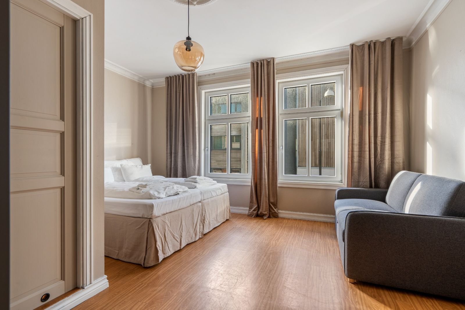 A bedroom with a bed, an armchair, large windows, beige curtains, and wooden flooring.