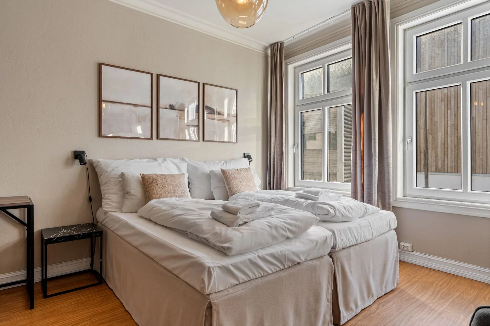 A neatly made bed with white bedding and two beige pillows in a sunlit bedroom. There are towel sets on the bed, three large windows with curtains, a small black marble-topped bedside table, and three framed pictures hanging above the bed.