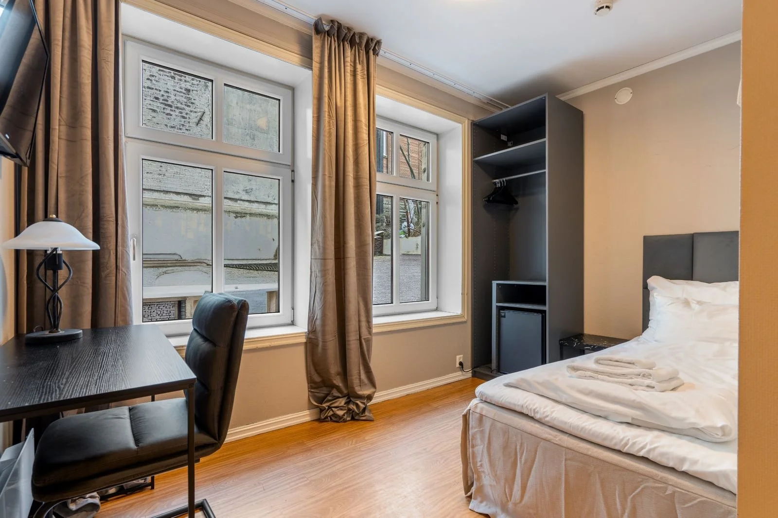 A small hotel room with a bed, a black desk with a lamp, a chair, and an open wardrobe. The room has two windows with beige curtains and views of an outdoor area.