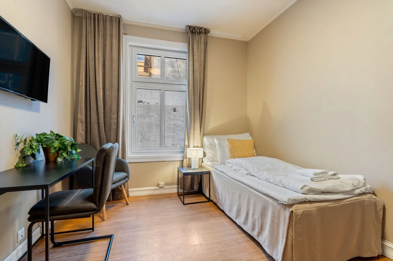A small, tidy hotel room with a single bed, a window with curtains, a small side table with a lamp, a chair, a desk with a potted plant, and a wall-mounted TV.