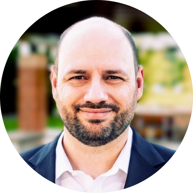 A smiling man with a beard and bald head, wearing a dark suit and white shirt, outdoors with blurred greenery in the background.