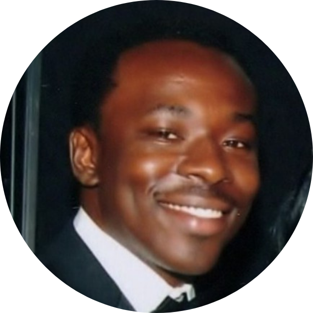 A young man with dark skin and short curly hair, smiling, wearing a black tuxedo with a white shirt, indoors at night.