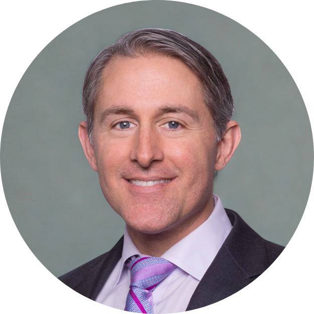 A professional headshot of a smiling man in a suit and tie with a grayish-green background.