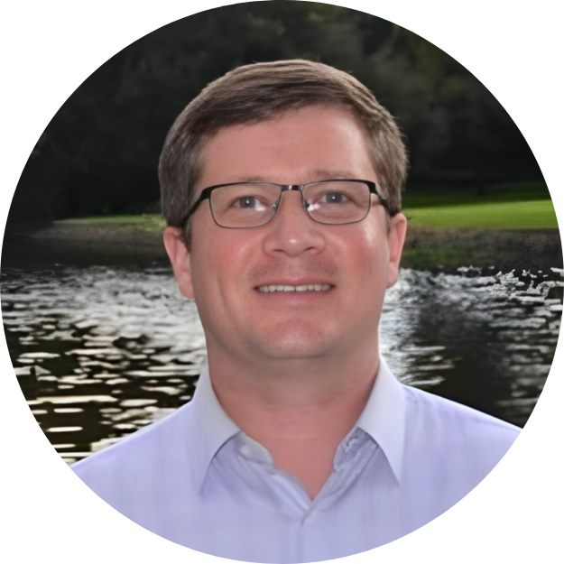 Portrait of a man with glasses wearing a light blue shirt outdoors near a body of water with green trees in the background.