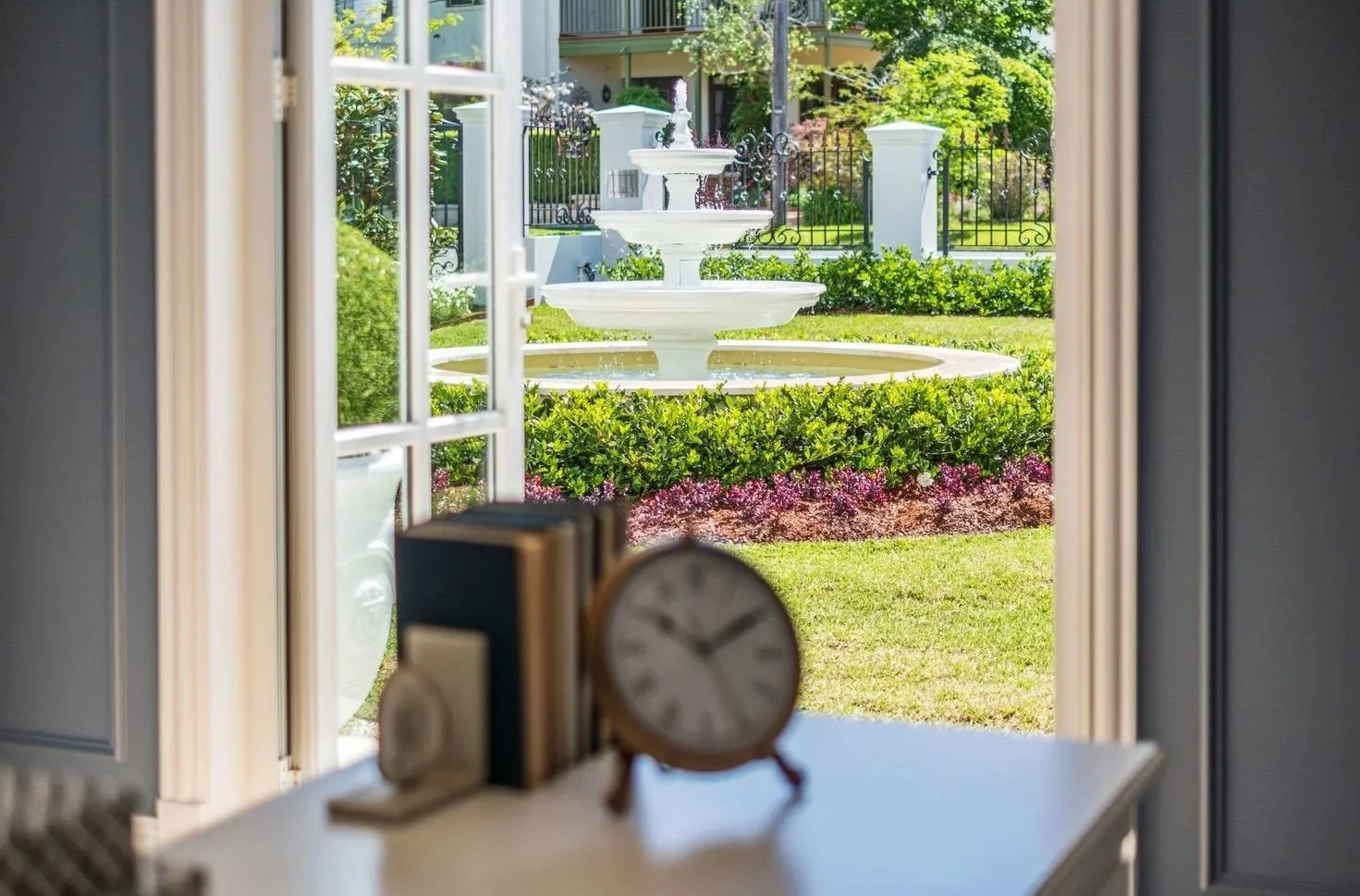 View of a white fountain with multiple tiers in a garden, seen through an open doorway inside a house. The garden has green grass, bushes, pink flowers, and a black iron fence in the background.