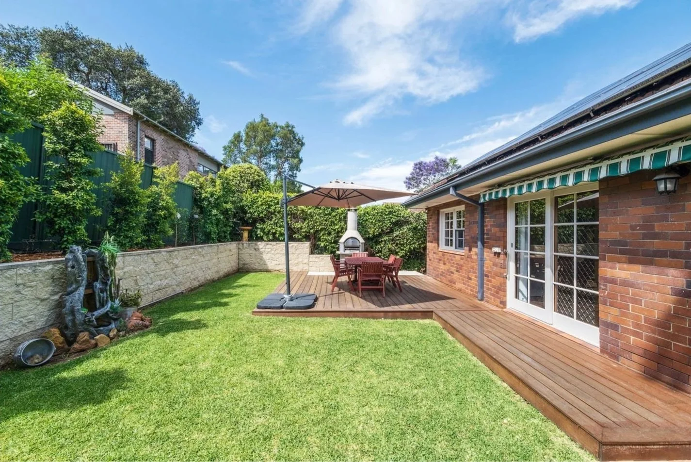 Backyard with green lawn, wooden deck, outdoor furniture, umbrella, brick house with sliding glass door, stone garden fountain, and lush bushes under a blue sky.