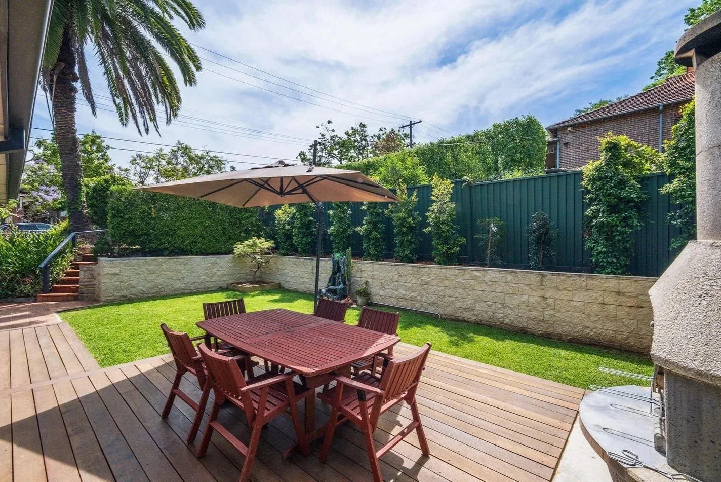 A backyard patio with a wooden table and six chairs under a large umbrella, a manicured lawn, a brick staircase, and a green fence with trees and shrubs behind it.