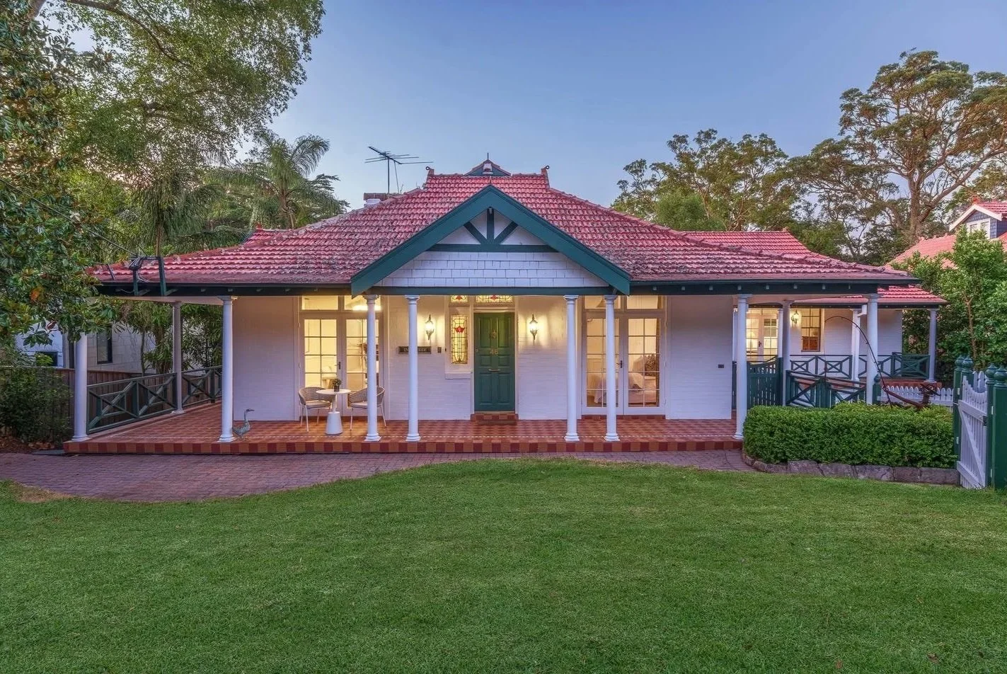 Front view of a cozy, single-story house with a red tiled roof, white walls, and a green front door. The house has a wide porch with white columns and outdoor seating, surrounded by a lush green lawn and trees.