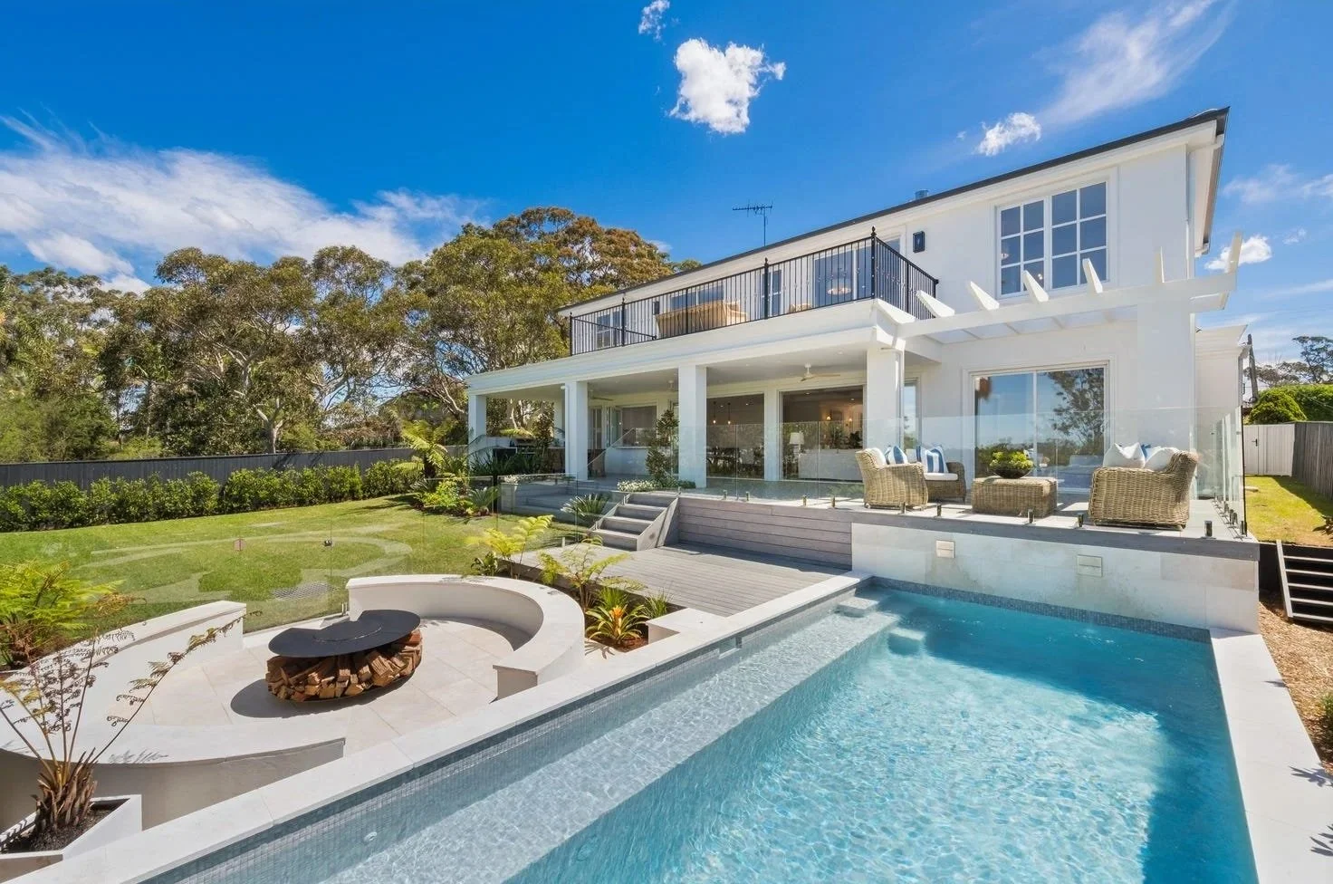 Modern two-story house with a large backyard, swimming pool, outdoor seating area with cushioned wicker chairs, glass railing, and a well-maintained lawn surrounded by trees under a blue sky with clouds.