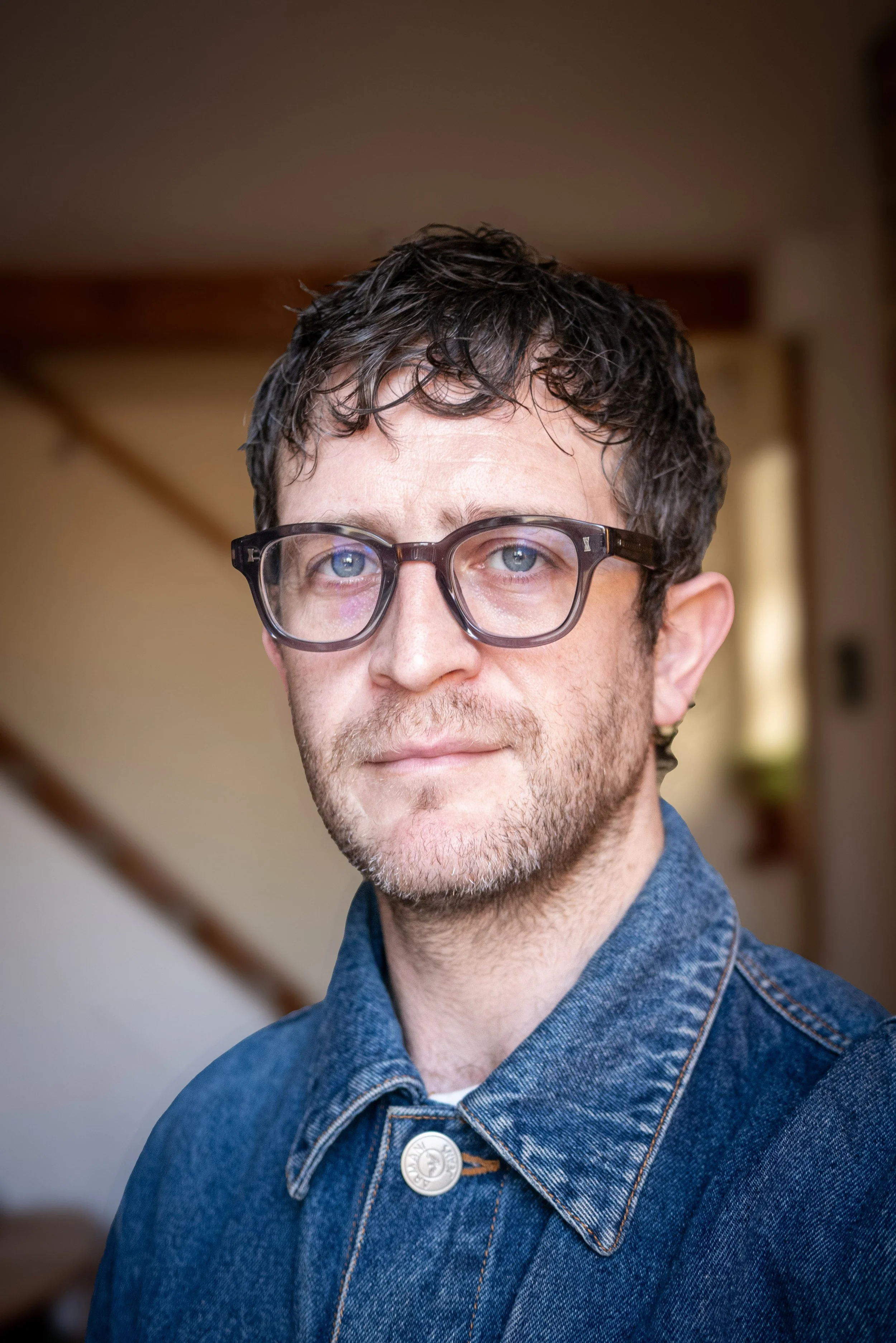 A man with glasses, short curly dark hair, and a light beard, wearing a denim jacket, looking at the camera indoors with a blurred background.