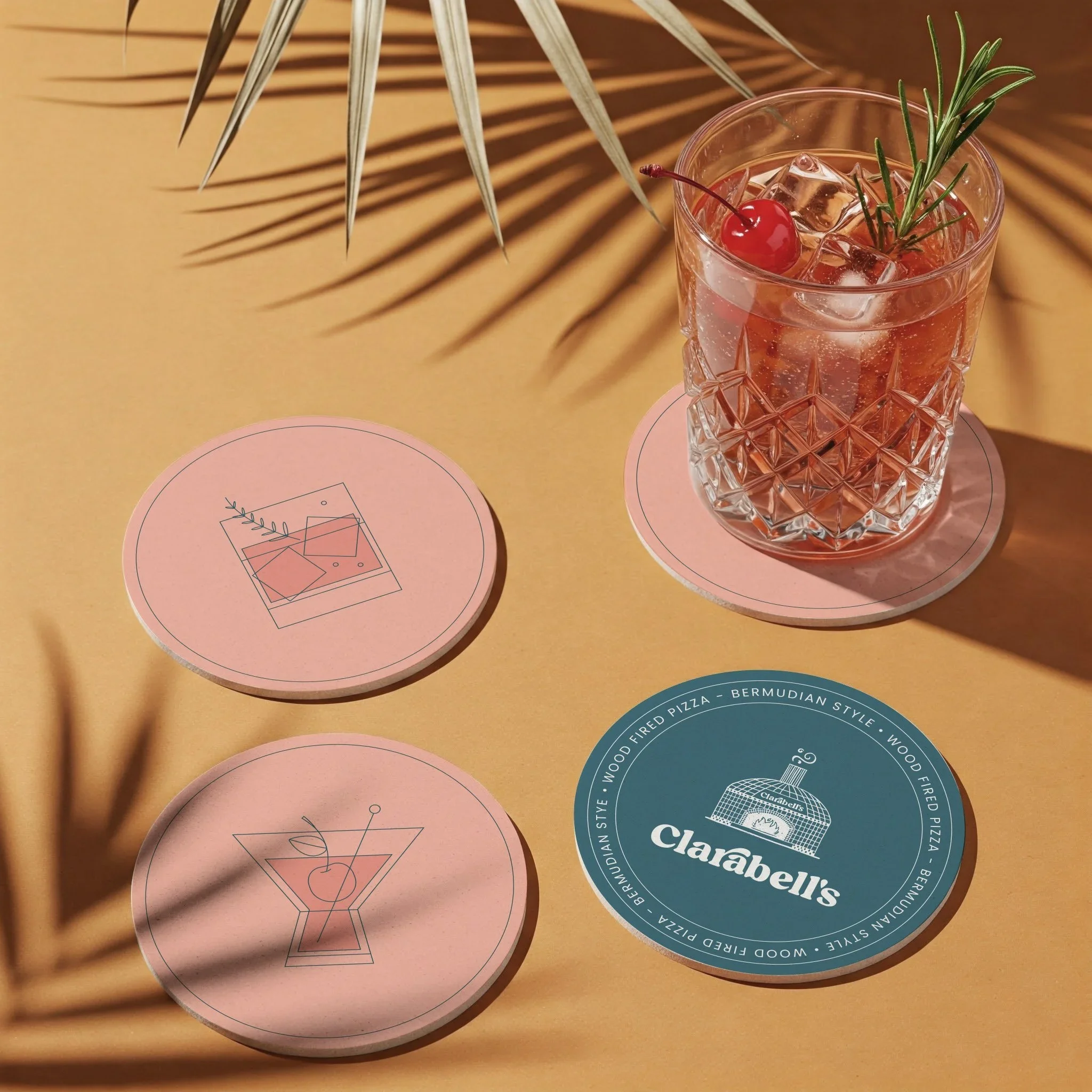 A glass of pink cocktail garnished with a cherry and rosemary sprig, placed on a pink coaster; two additional pink coasters with abstract line art designs, and a blue coaster with a bakery logo and text, all on a yellow surface with shadowed palm frond.
