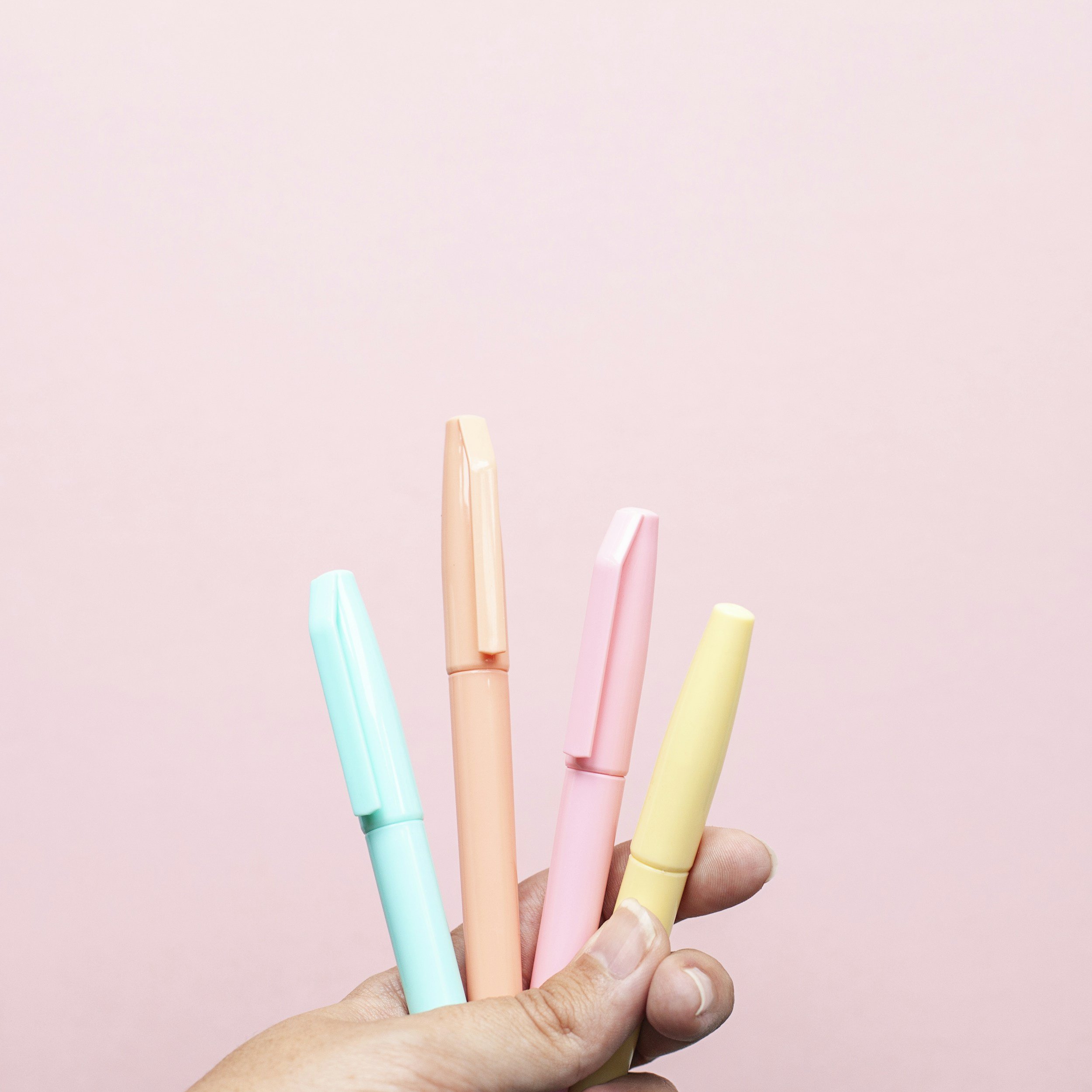 Hand holding four pastel-colored pens against a pink background.