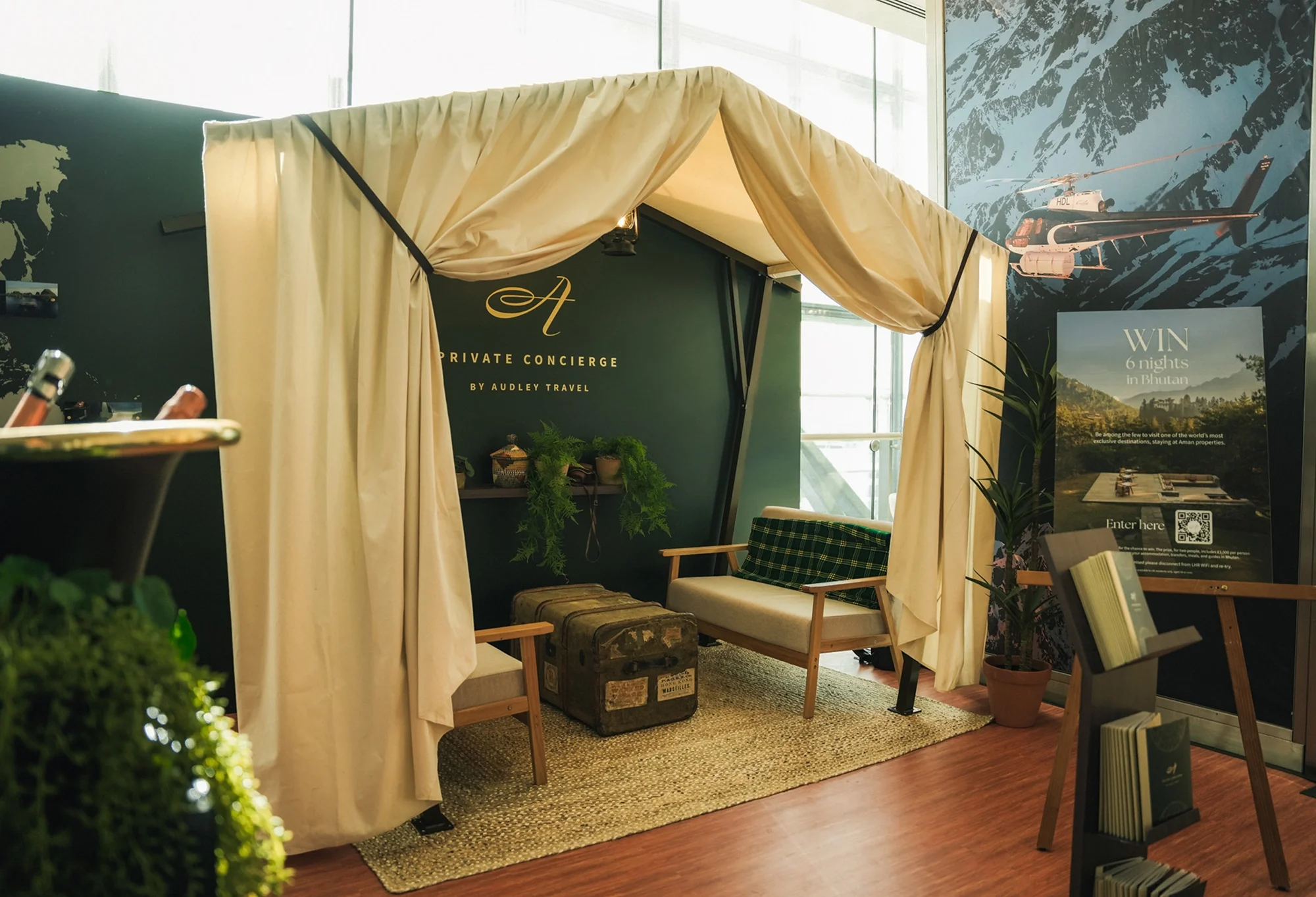 A private concierge booth with cream curtains, a green wall, and woodland decor, including travel-themed posters and seating for visitors.