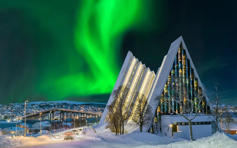 Snow-covered modern church with glass windows and sharp angles, illuminated at night, against the backdrop of the Northern Lights and a city in winter.