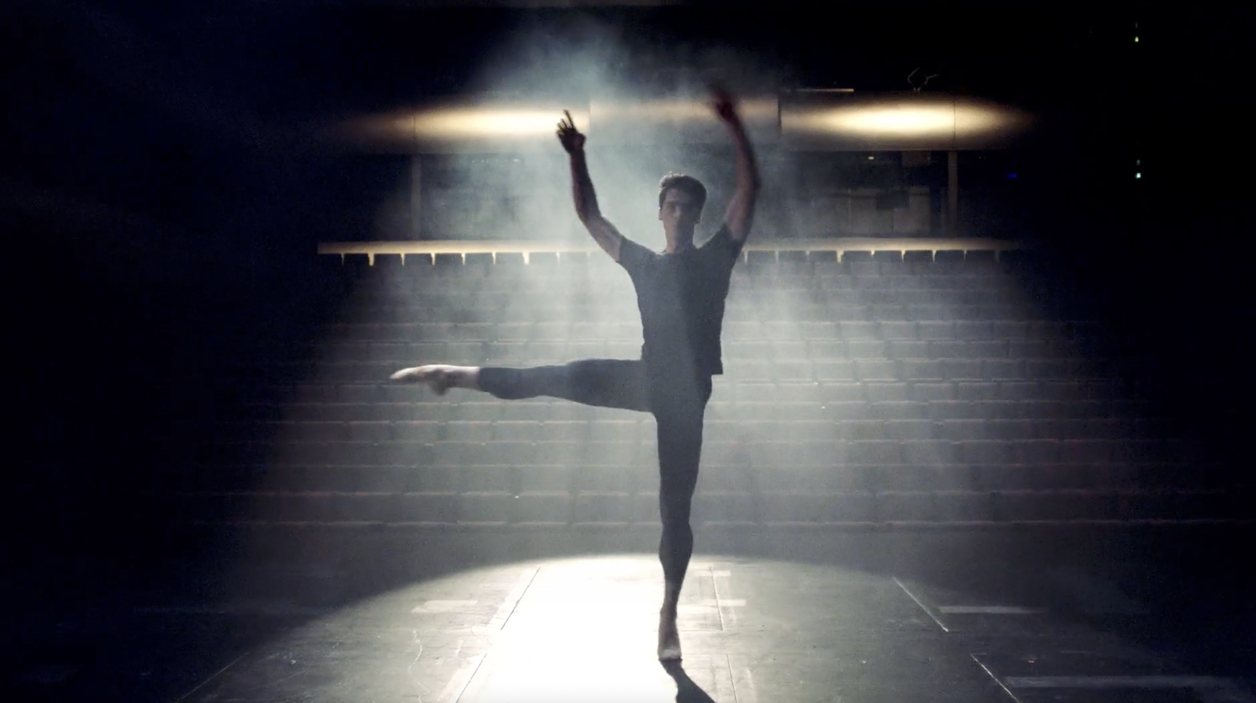 A male ballet dancer practicing alone on a dimly lit stage, with spotlight and smoke creating a dramatic atmosphere.
