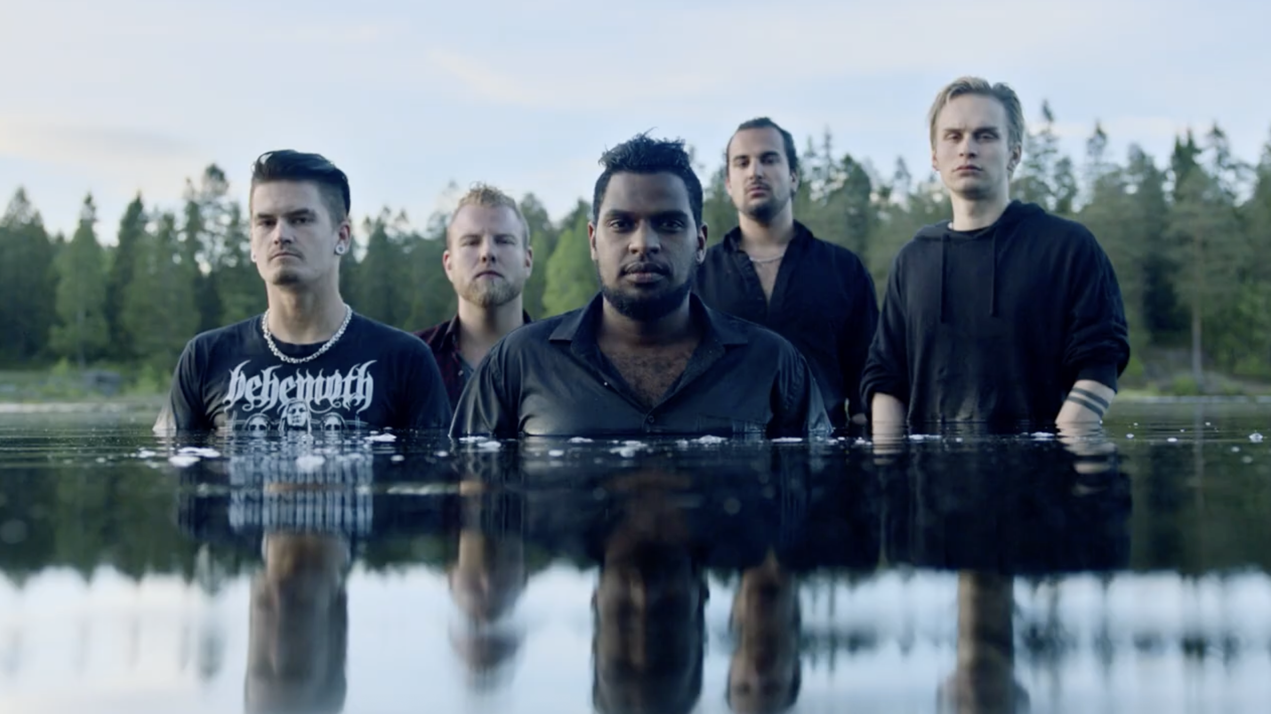 Five men standing in water outdoors with forest and sky in the background.