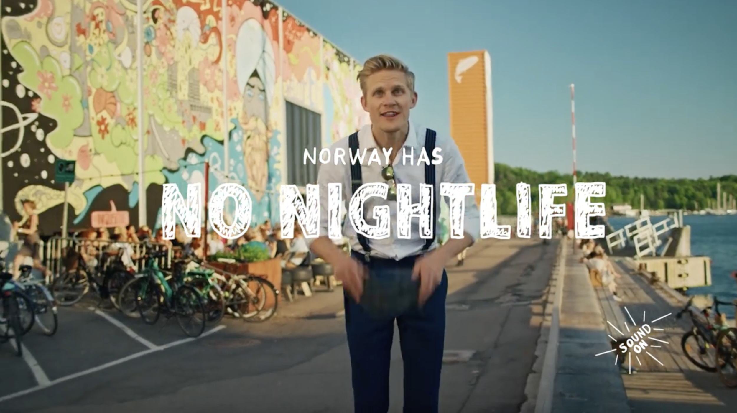 A young man standing on a waterfront promenade in Norway, with a colorful mural on a building behind him, bicycles parked nearby, and the sea in the background. The text overlay reads 'Norway Has No Nightlife' and 'Sound On'.