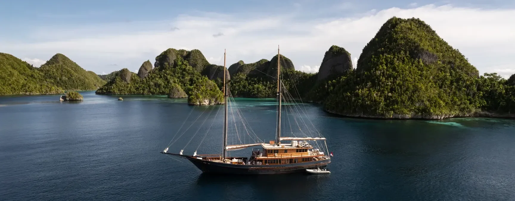 A large black sailing yacht with two tall masts floating on calm water surrounded by green, rocky islands in a tropical area.