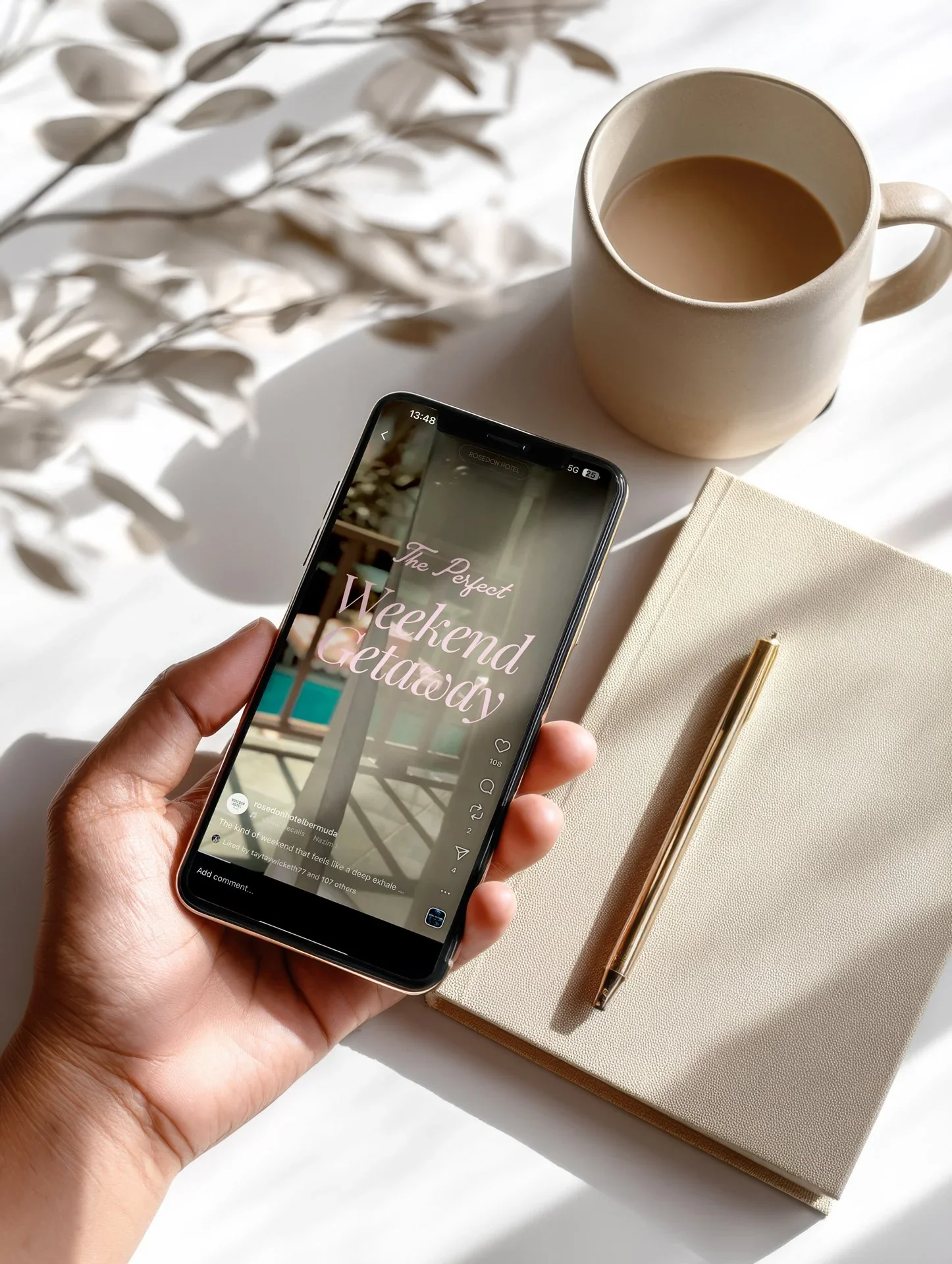 A person holding a smartphone displaying a social media post that says 'The Perfect Weekend Getaway' next to a beige notebook with a gold pen, a cup of coffee or tea, and some leafy plant shadows in the background.