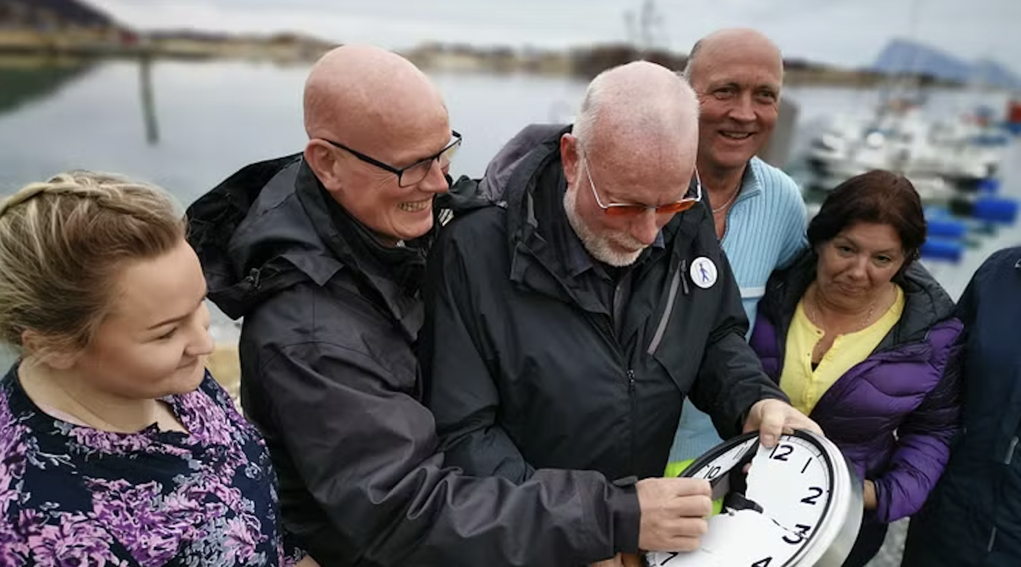 Group of six people outdoors by water, gathered around a clock, with one person adjusting the time.