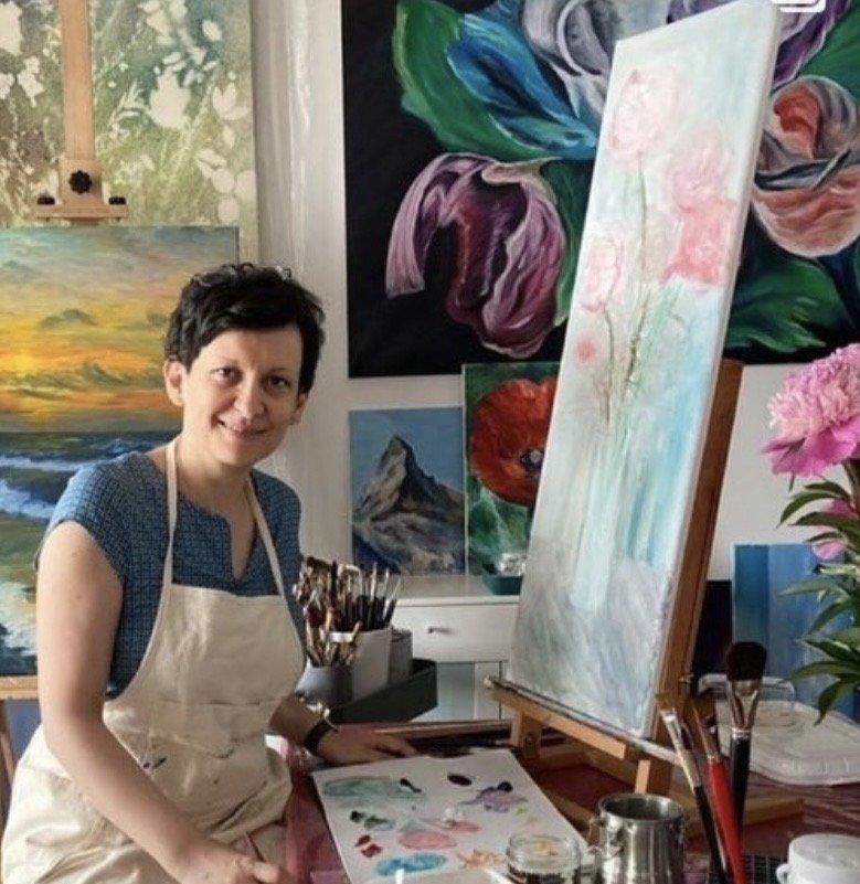 Female artist painting in her studio, surrounded by canvases including a large pastel floral artwork and landscape paintings in the background.