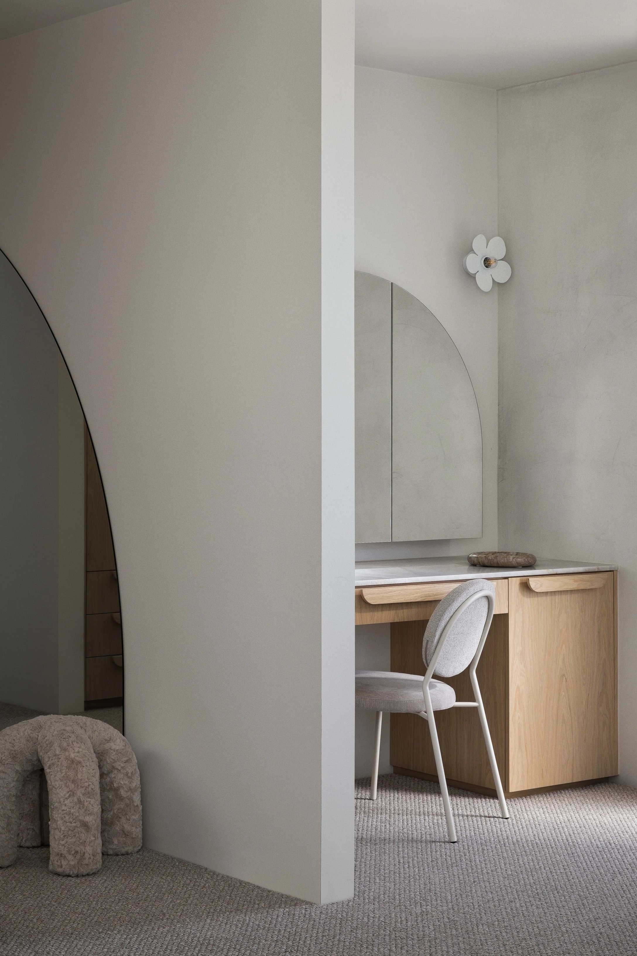 Minimalist bedroom corner with a vanity table, mirror, cushioned chair, and a decorative flower wall lamp.