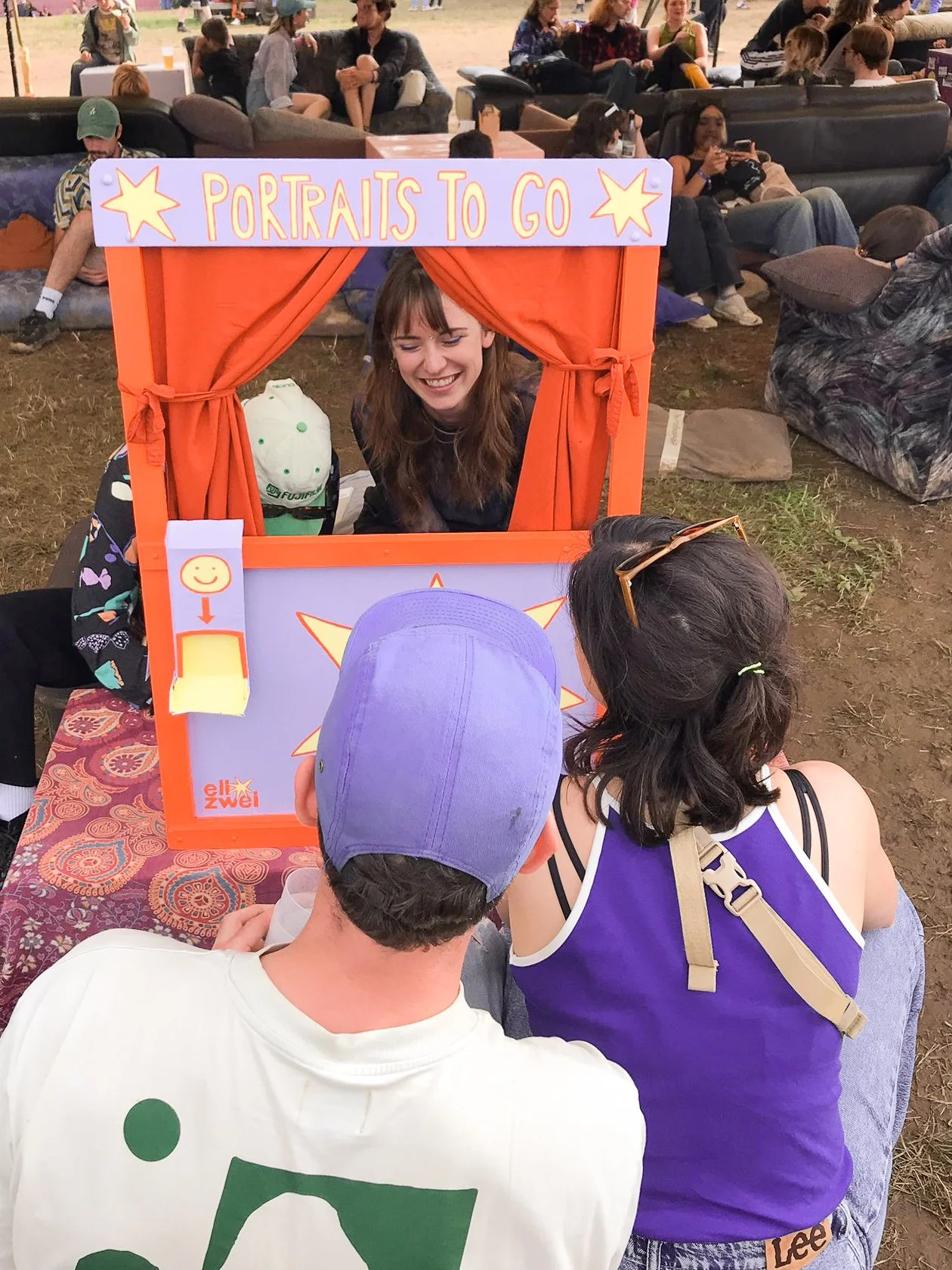 Zwei Personen sitzen vor einer live Zeichen-Station mit  dem Text 'Portraits to go' von ellmalzwei. Dahinter sitzen zwei Frauen, die lachen. Umgeben von mehreren Menschen auf einem Festival.