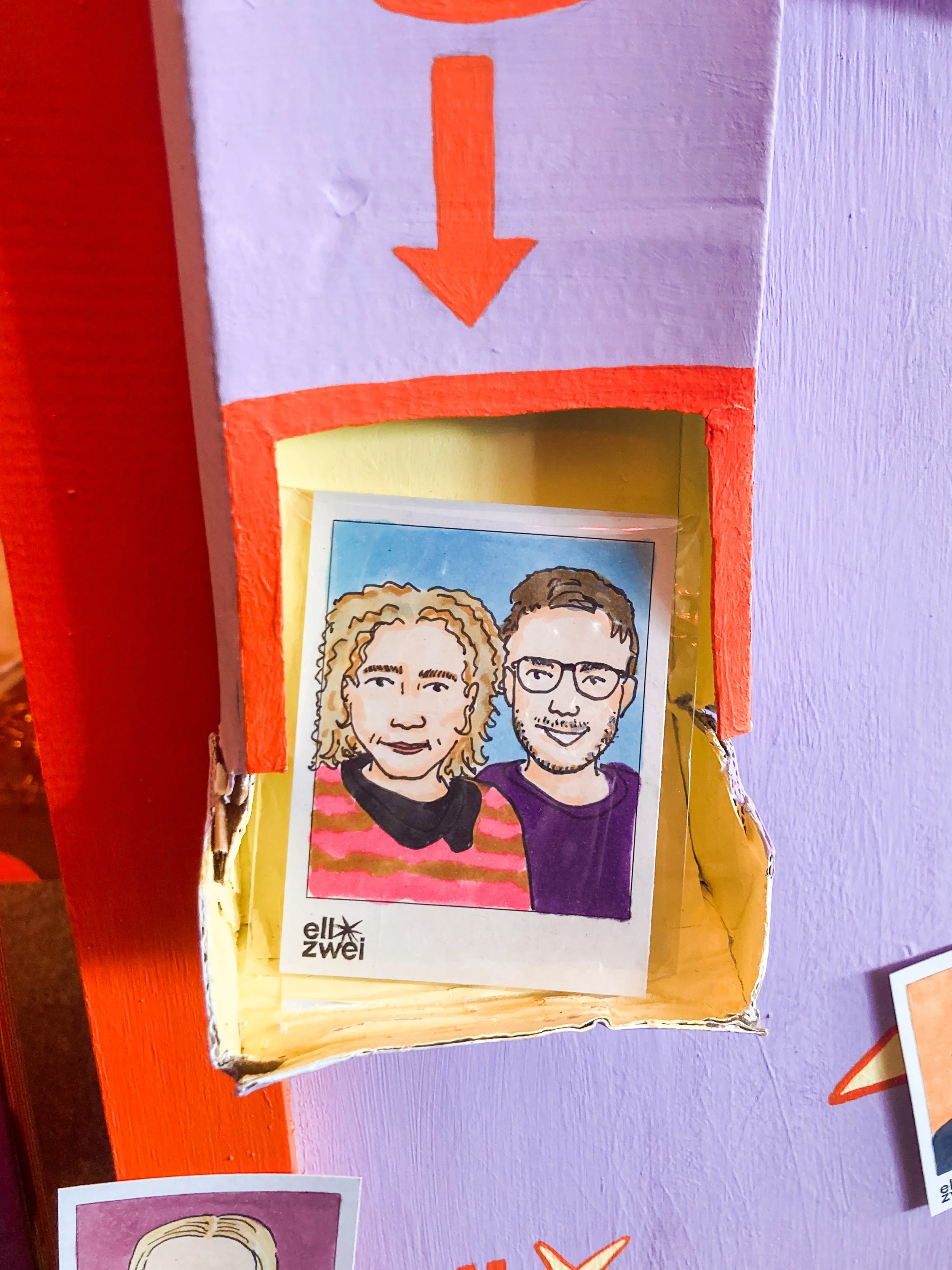 Ein handbemalter Fotoauswurf mit einem roten Pfeil nach unten darüber. Darin befindet sich ein handgezeichnetes Portrait von zwei Personen, einem Mann und einer Frau.