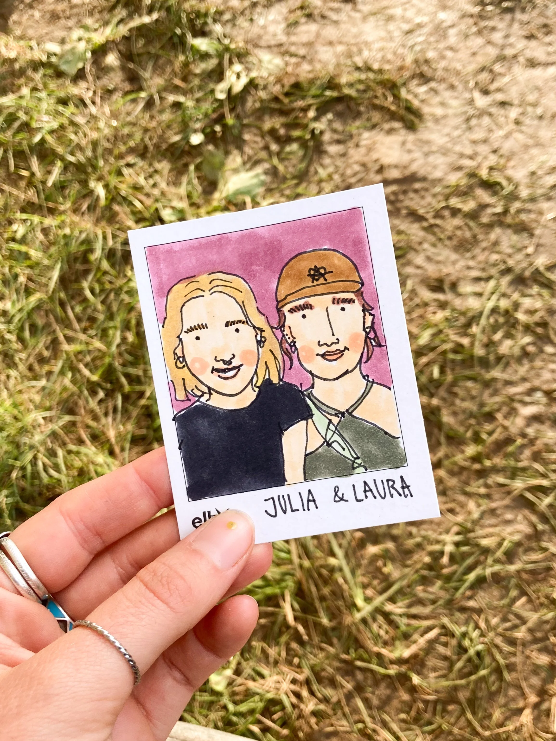 Eine hand hält eine kleine Illustration von zwei lachenden Frauen, Julia und Laura, vor einem Naturhintergrund mit Gras und Erde.