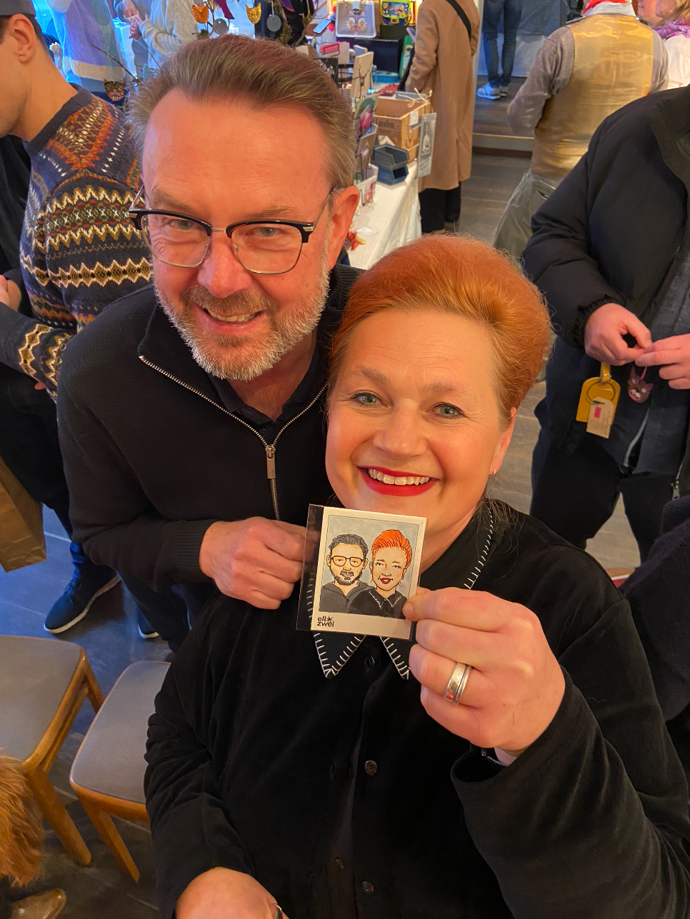 Zwei Menschen, ein Mann mit Brille und grauem Bart, und eine Frau mit roten Haaren, lachen in einem Laden. Die Frau hält ein handgezeichnetes Portraitbild von ihnen in der Hand.