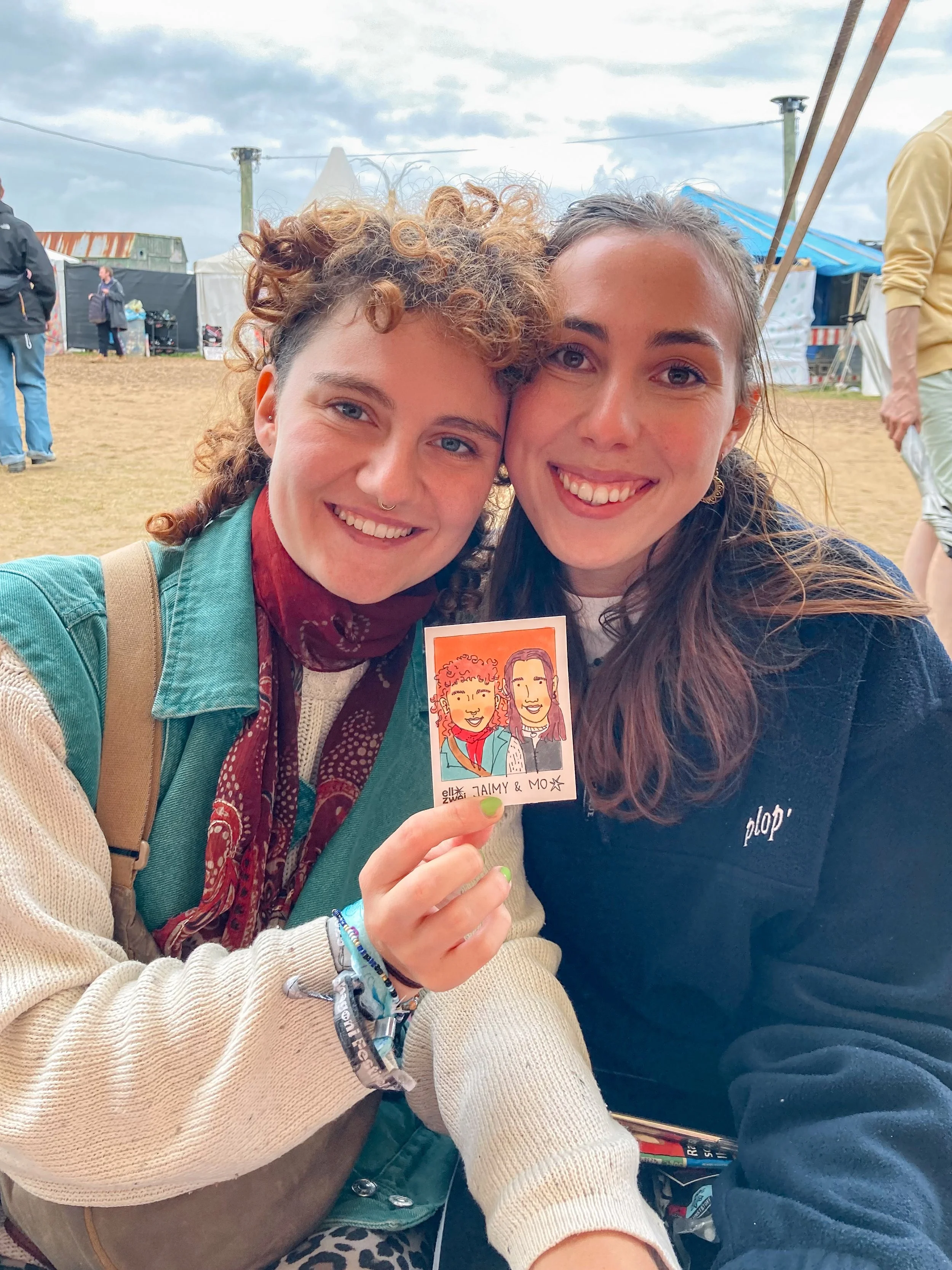 Zwei junge Frauen lachen und halten ein handgezeichnetes Portraitbild der beiden bei einem Outdoor-Festival. Im Hintergrund sind Zelte und Menschen zu sehen.