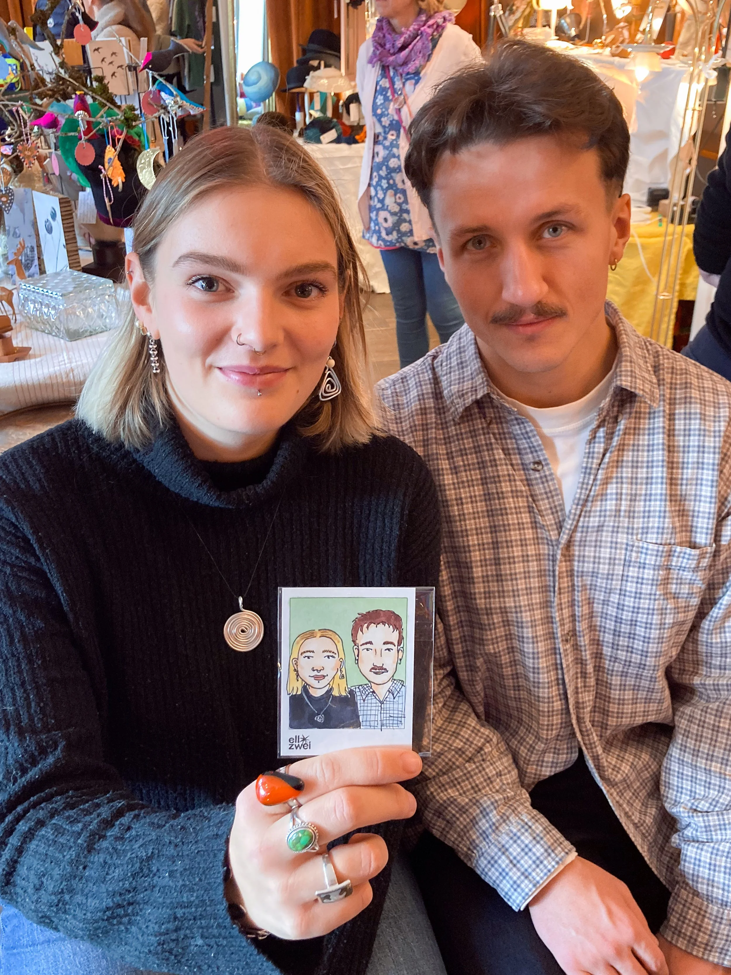 Ein junges Paar sitzt nebeneinander auf einer Veranstaltung, die Frau zeigt ein handgezeichnetes Portraitbild der beiden, im Hintergrund sind Stände mit Dekorationen und anderen Menschen zu sehen.