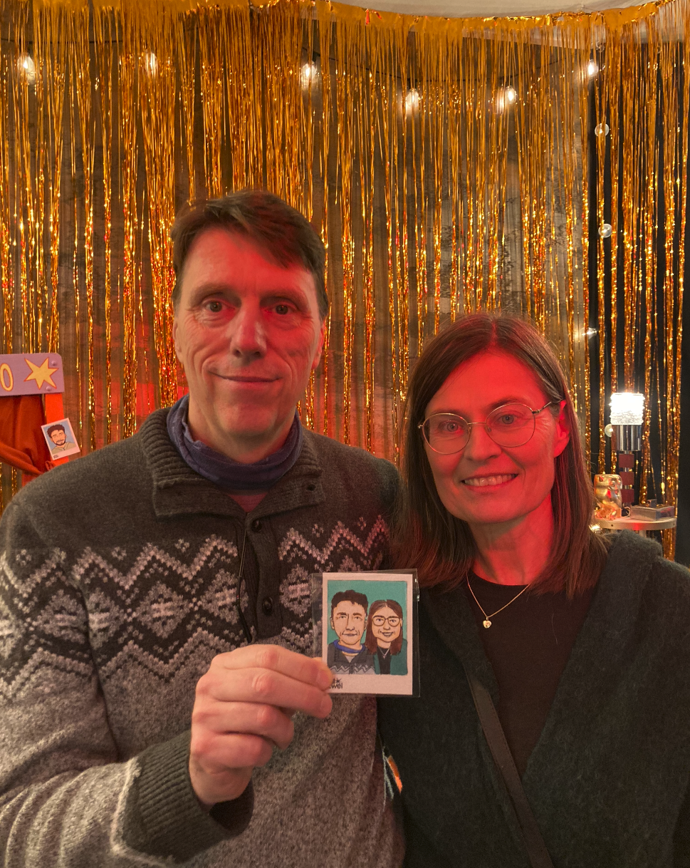Ein Mann und eine Frau stehen vor einem goldfarbenen Lametta Vorhang und posieren für ein Foto. Der Mann hält ein kleines handgezeichnetes Porträtbild der beiden Personen in der Hand.
