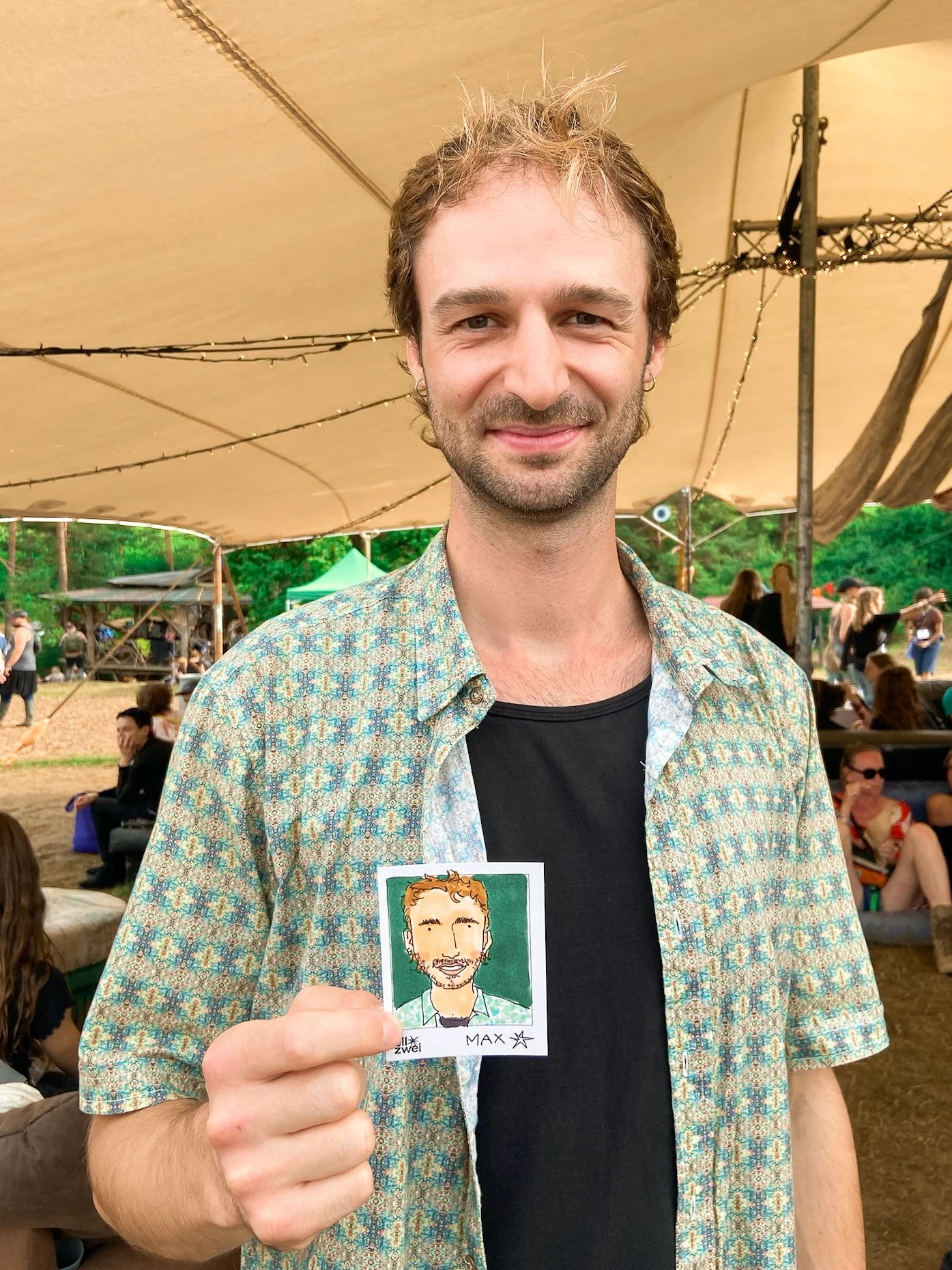 Ein Mann auf einem Festival hält eine karikaturartige Zeichnung von sich selbst in der Hand, vor einem Zelt mit anderen Besuchern.