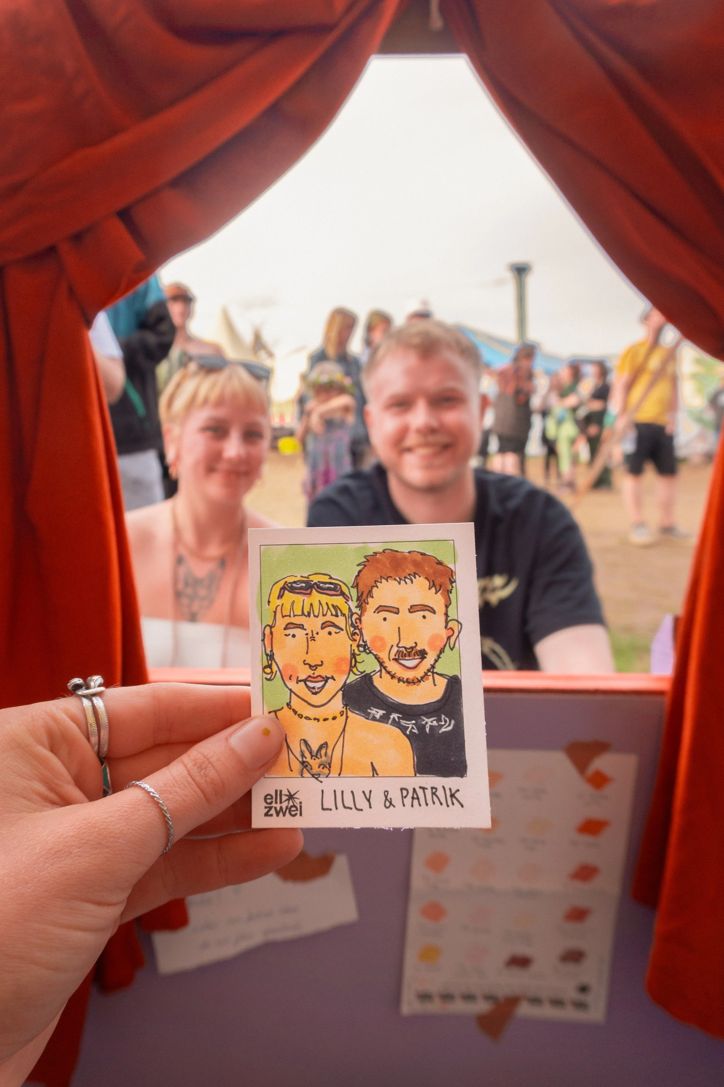 Eine Person hält eine handgezeichnete Karikatur von Lilly und Patrik in der Hand, während dahinter zwei Menschen auf einem Festival vor einem offenen, roten Vorhang  einer Portrait-Station sitzen und lächeln.
