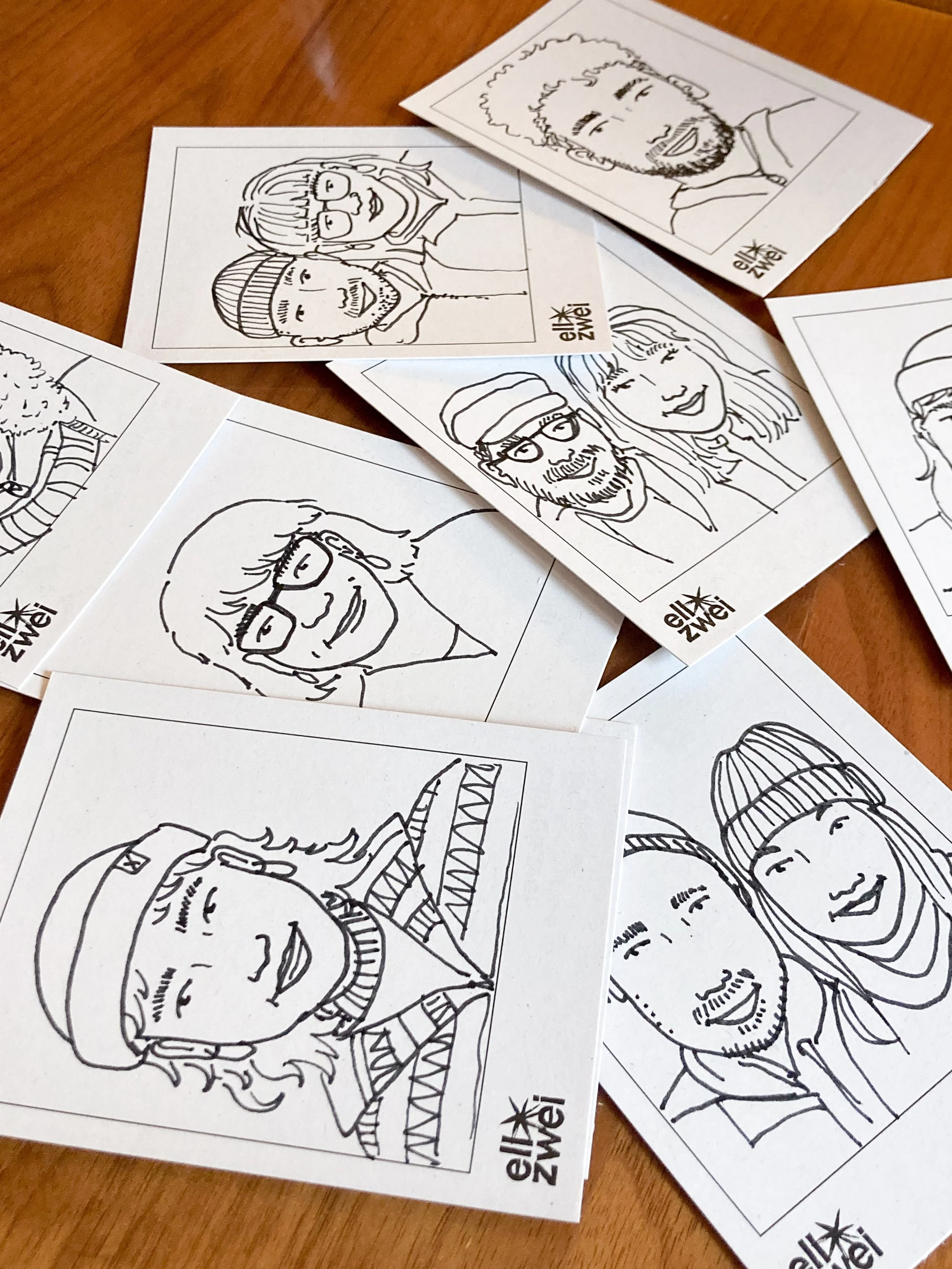 Mehrere kleine Portrait-Zeichnungen von Menschen auf Papier, die auf einem Holztisch liegen. Die Zeichnungen sind Strichzeichnungen und zeigen verschiedene Personen, einige lächelnd, andere lässig. Einige tragen Brillen, Mützen oder Hüte.