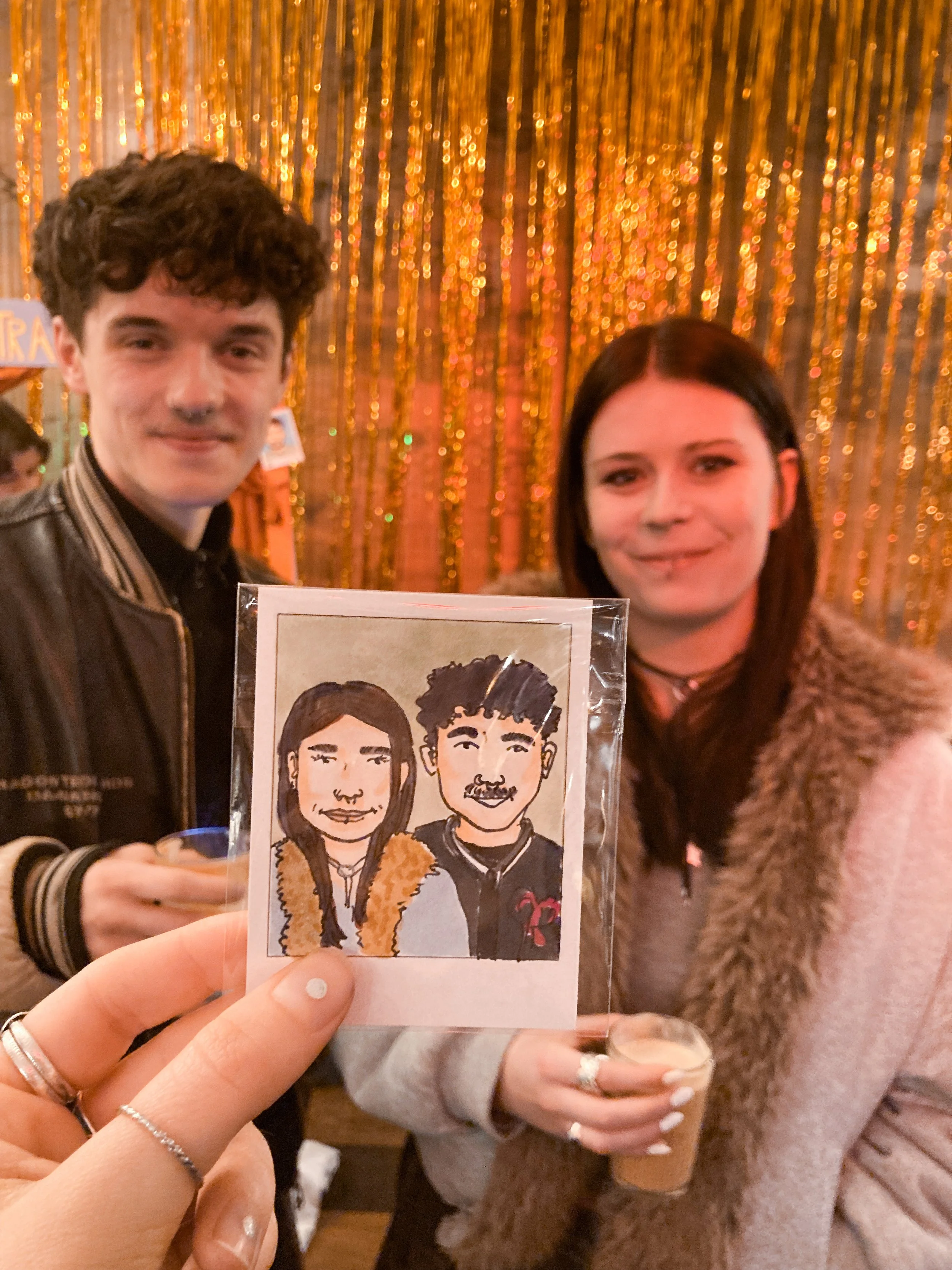 Zwei Personen stehen vor einer goldfarbenen, glänzenden Dekoration und halten ein handgezeichnetes Porträt in einer transparenten Hülle. Die linke Person hat braune, lockige Haare, trägt eine schwarze Lederjacke. Die rechte Person hat lange, dunkle H
