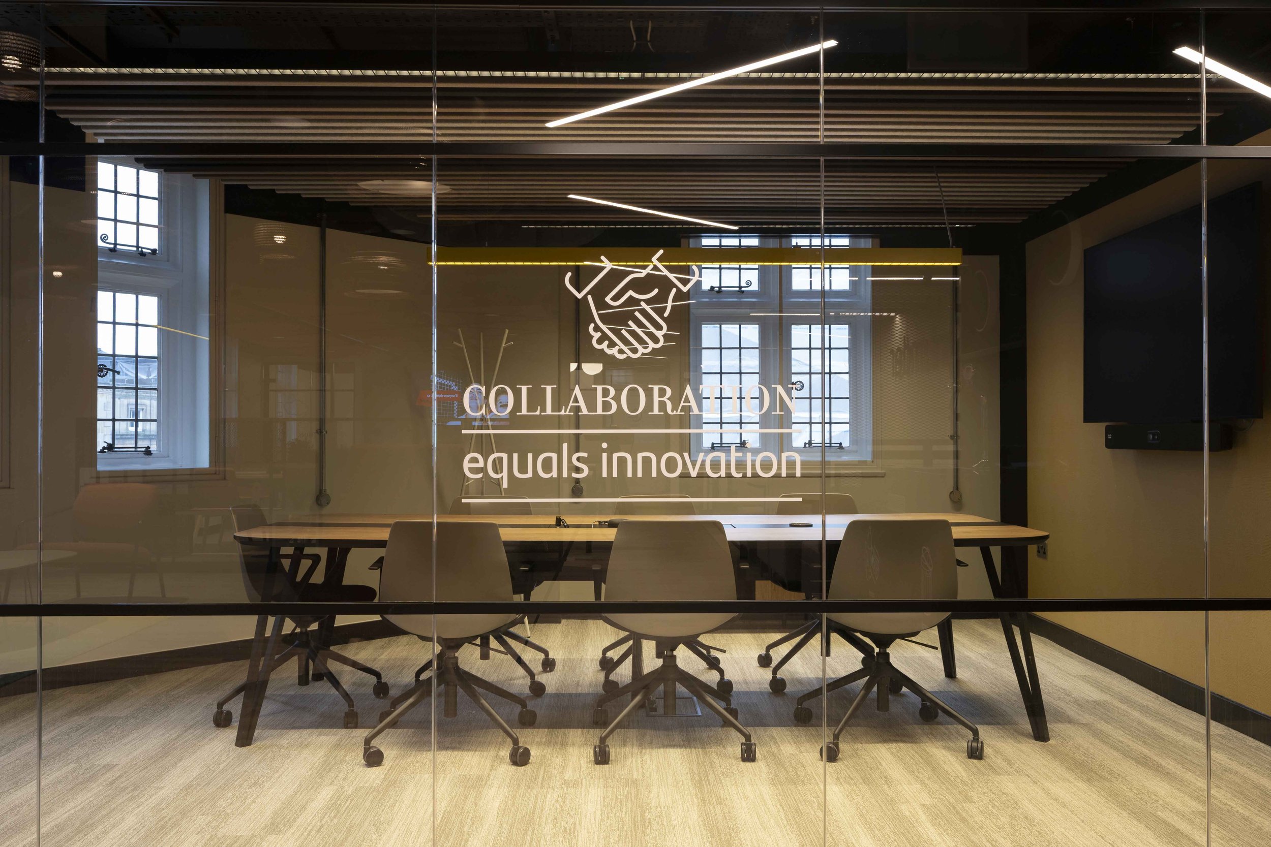 Modern conference room with glass walls, wooden ceiling, rows of chairs and tables, and a neon sign of a handshake with the words 'COLLABORATION equals innovation'.