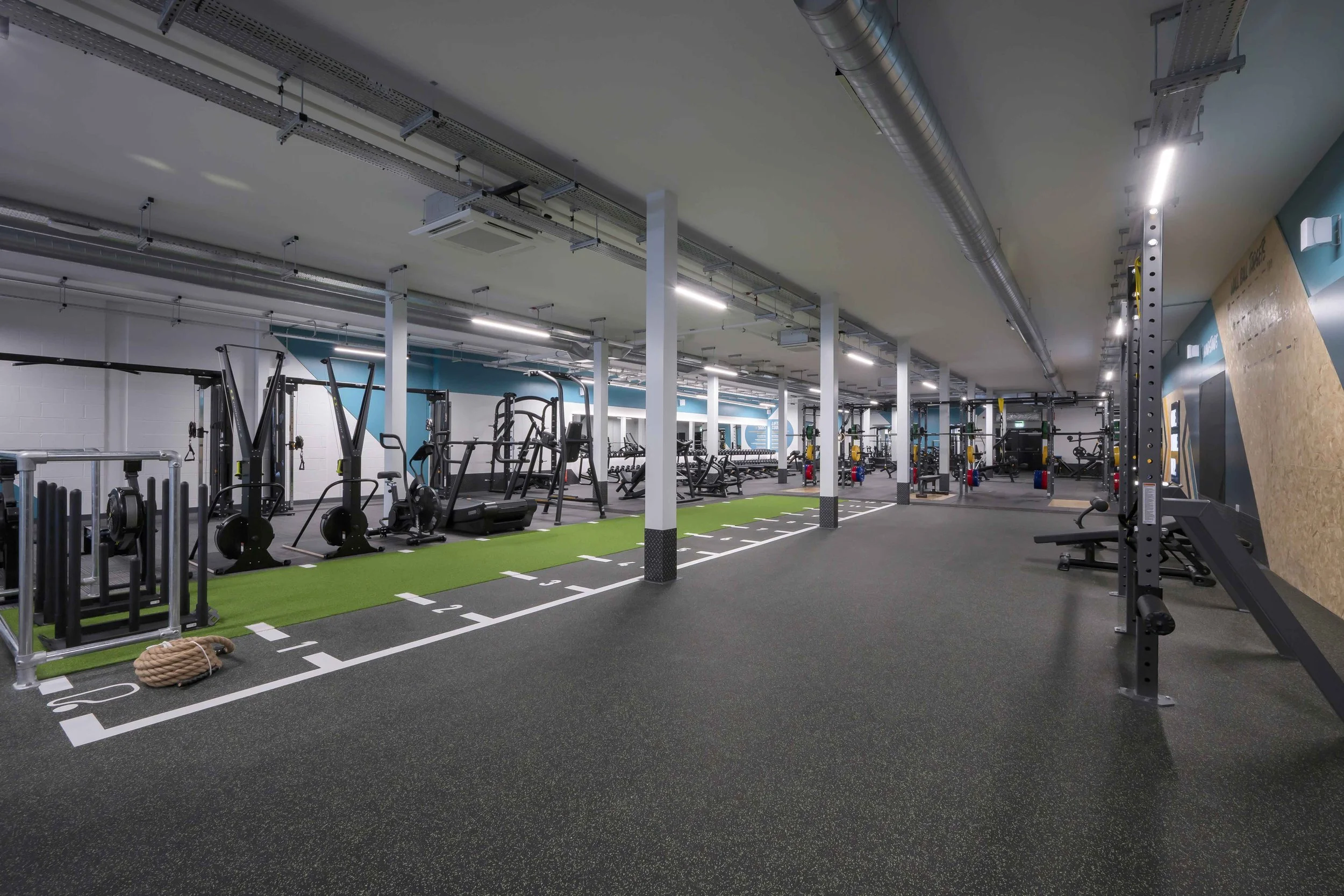 Empty gym with weightlifting equipment, treadmills, and a rock climbing wall, with black rubber flooring and overhead lighting.