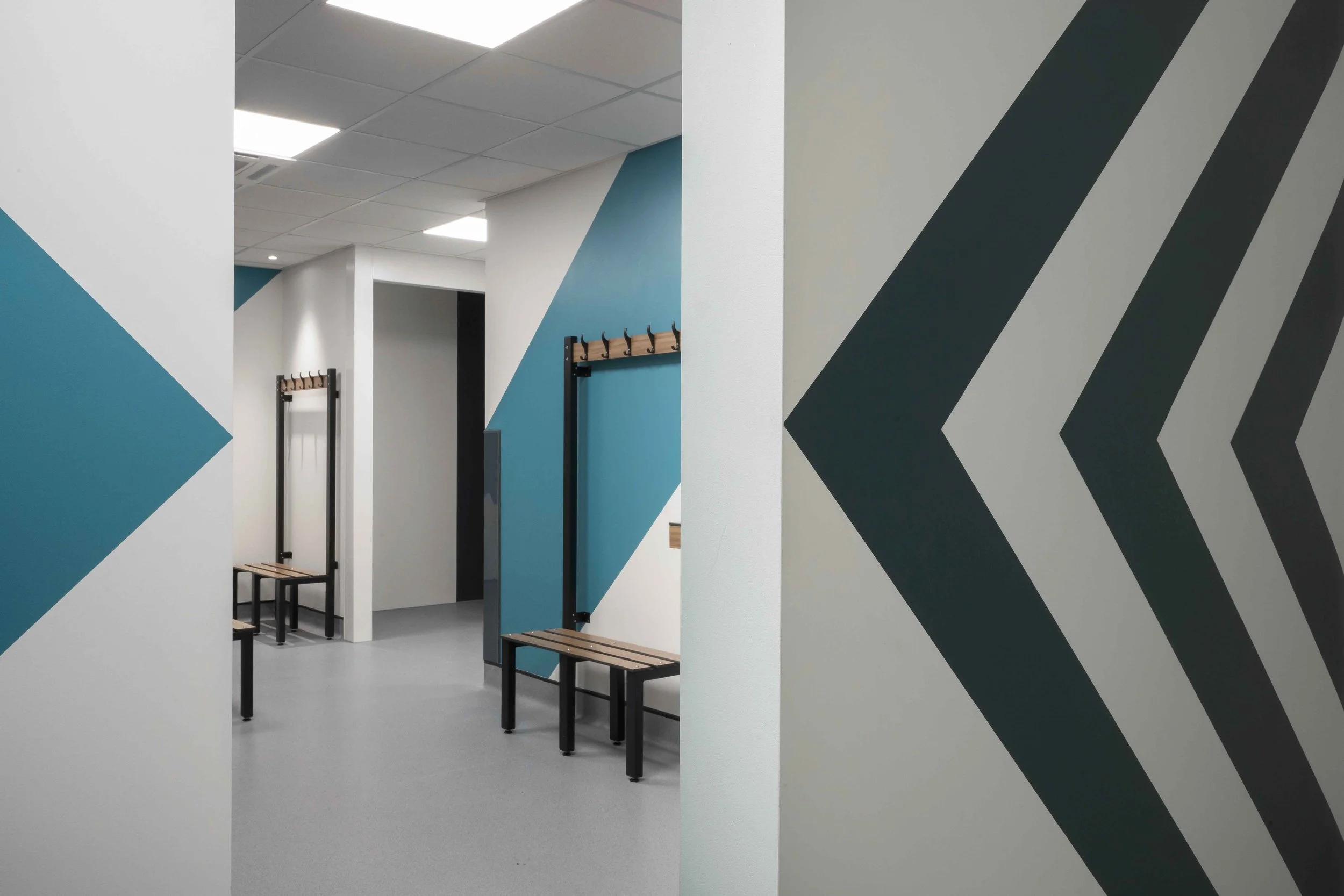 Interior of a modern office or waiting area with walls painted in geometric patterns of teal, white, and dark gray. There are wooden benches and hooks on black frames along the walls, and a doorway can be seen in the background. The ceiling has white