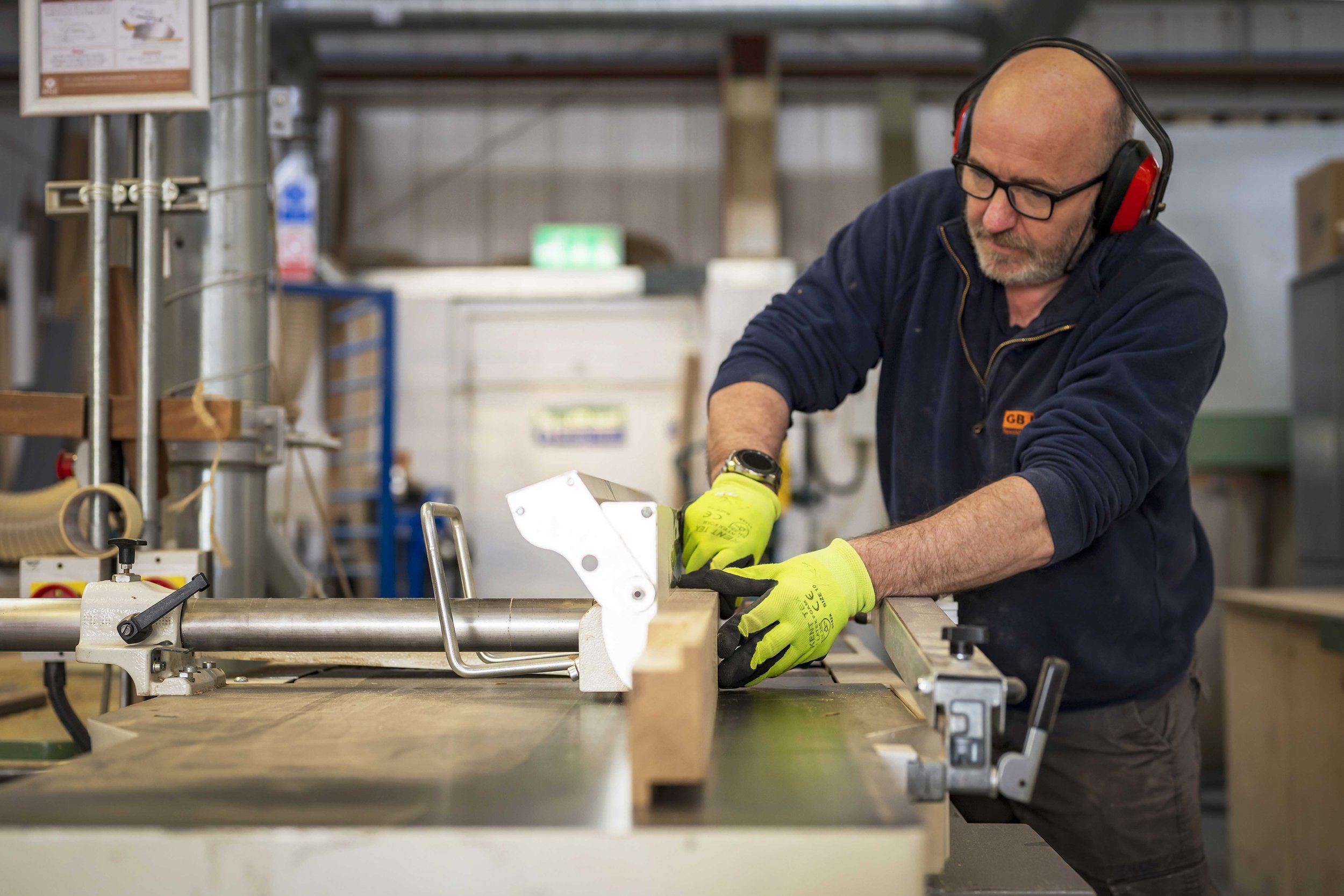 A man working in a workshop, wearing safety glasses, yellow gloves, and earmuffs, handling a metal piece on a workbench.