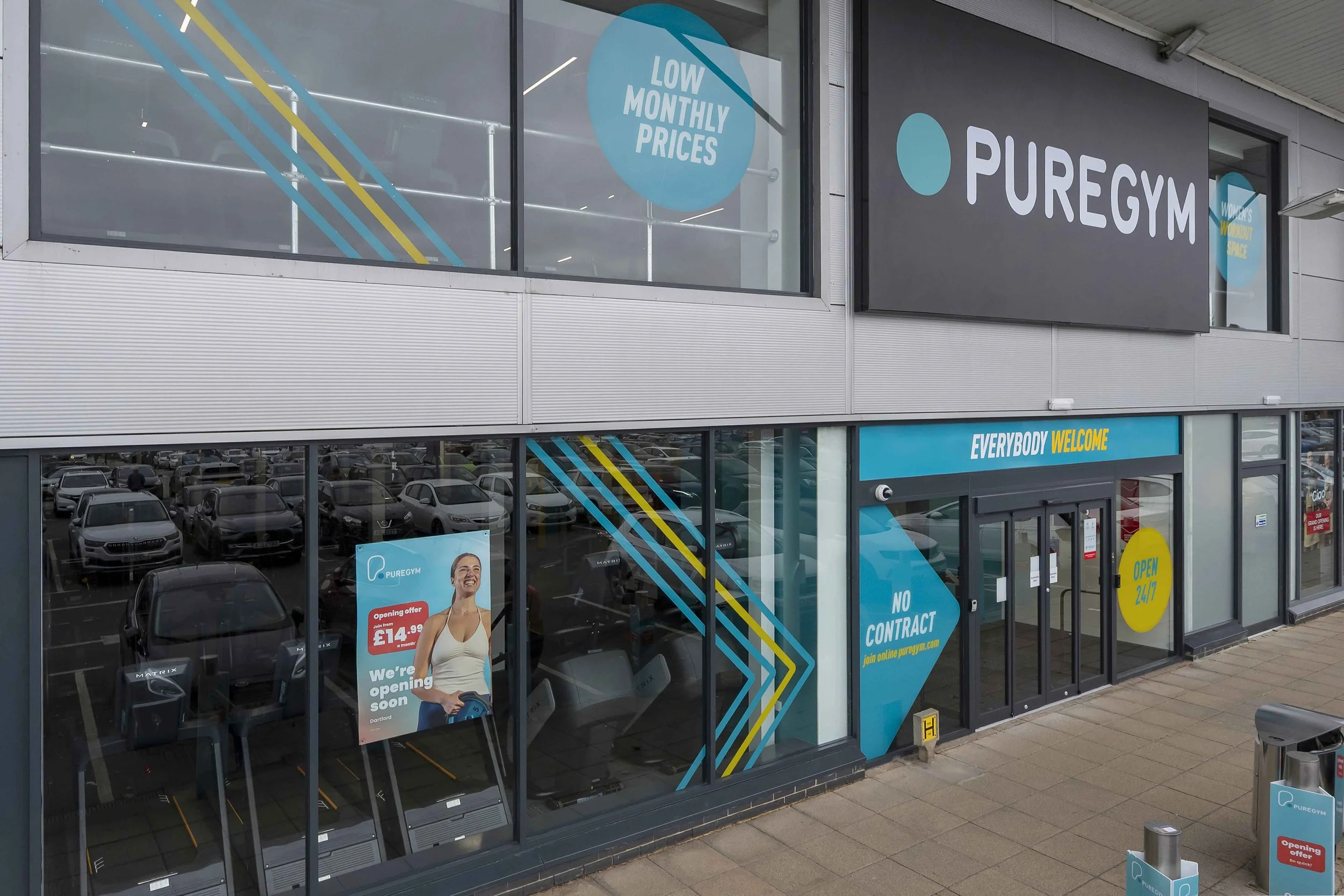The storefront of PureGym, a fitness center, with large windows displaying signs that say 'Low Monthly Prices' and 'Everybody Welcome.' The entrance has a sign indicating it is open 24/7 and that there is no contract required.