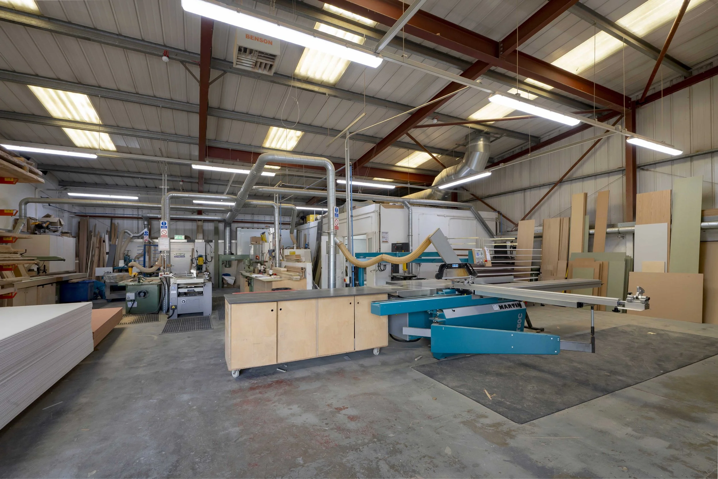 A woodworking workshop with machinery, workbenches, and stacks of wood panels. Overhead fluorescent lights illuminate the space, which has a metal roof and beams.