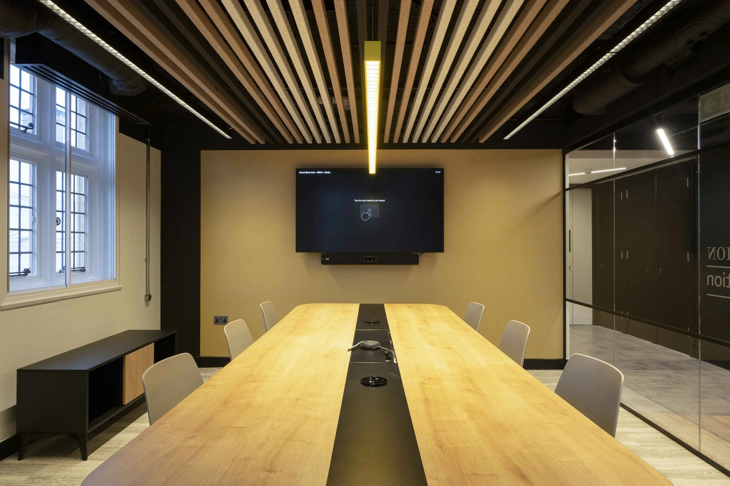 Modern conference room with a large wooden table, white chairs, a black wall-mounted screen, large windows on the left, and a glass wall on the right.