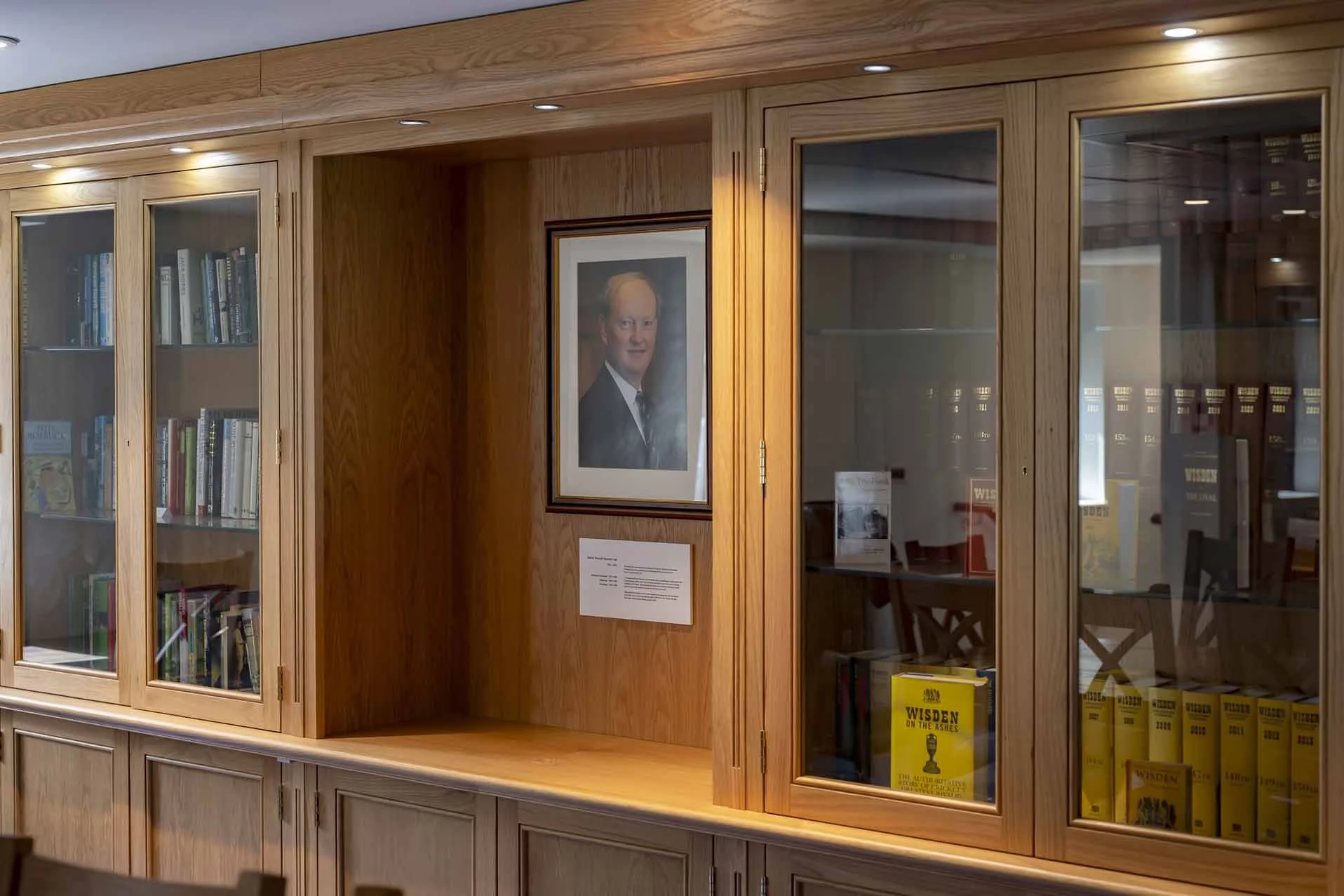 A wooden display cabinet with glass doors filled with books, including some titled 'Wisden on the Ashes,' and a framed photograph of a man hanging on the wall beside it.