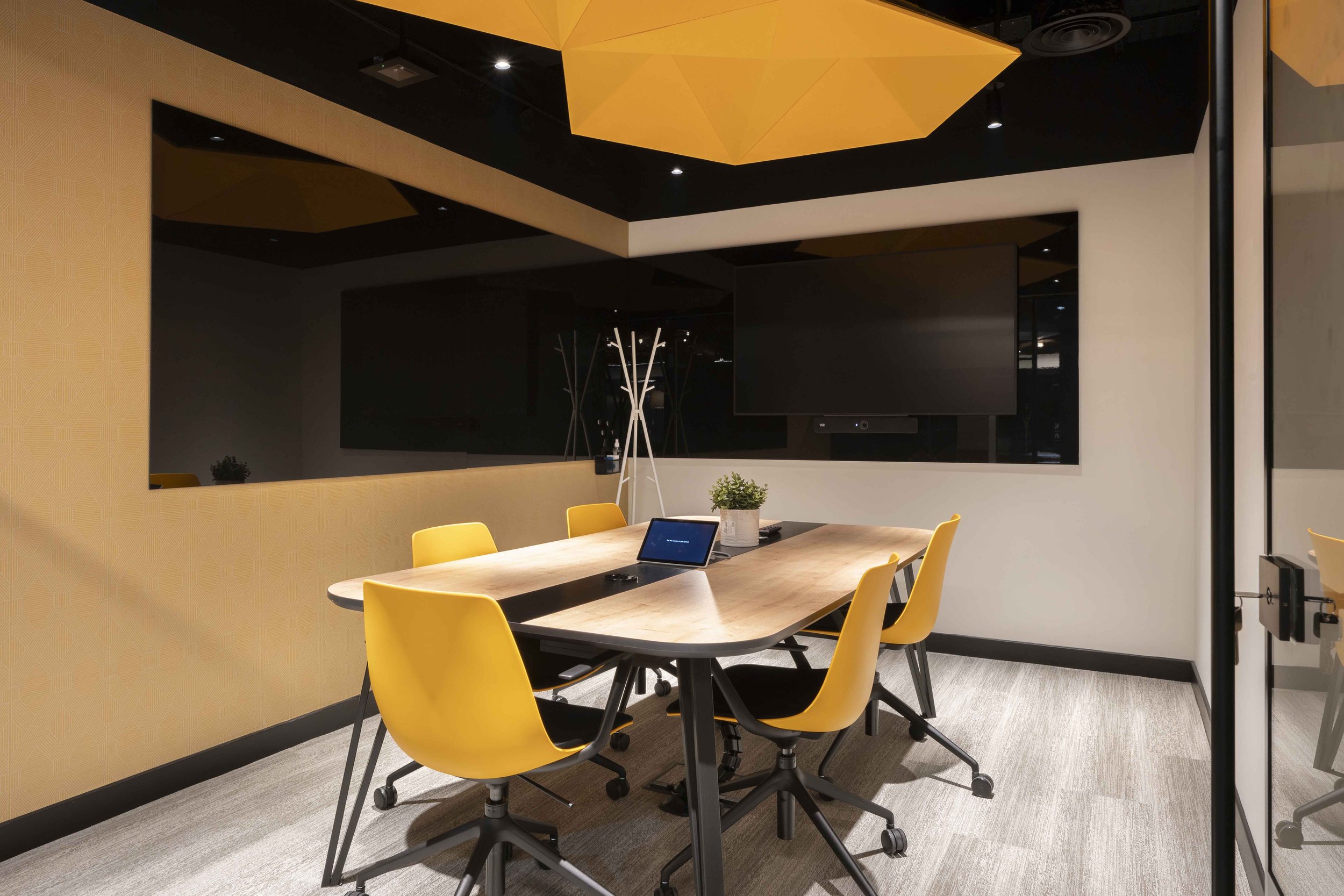 A modern conference room with a long wooden table surrounded by yellow chairs. A large black television screen is mounted on the wall, and decorative plant arrangements are placed on the table. The ceiling features a yellow geometric light fixture.