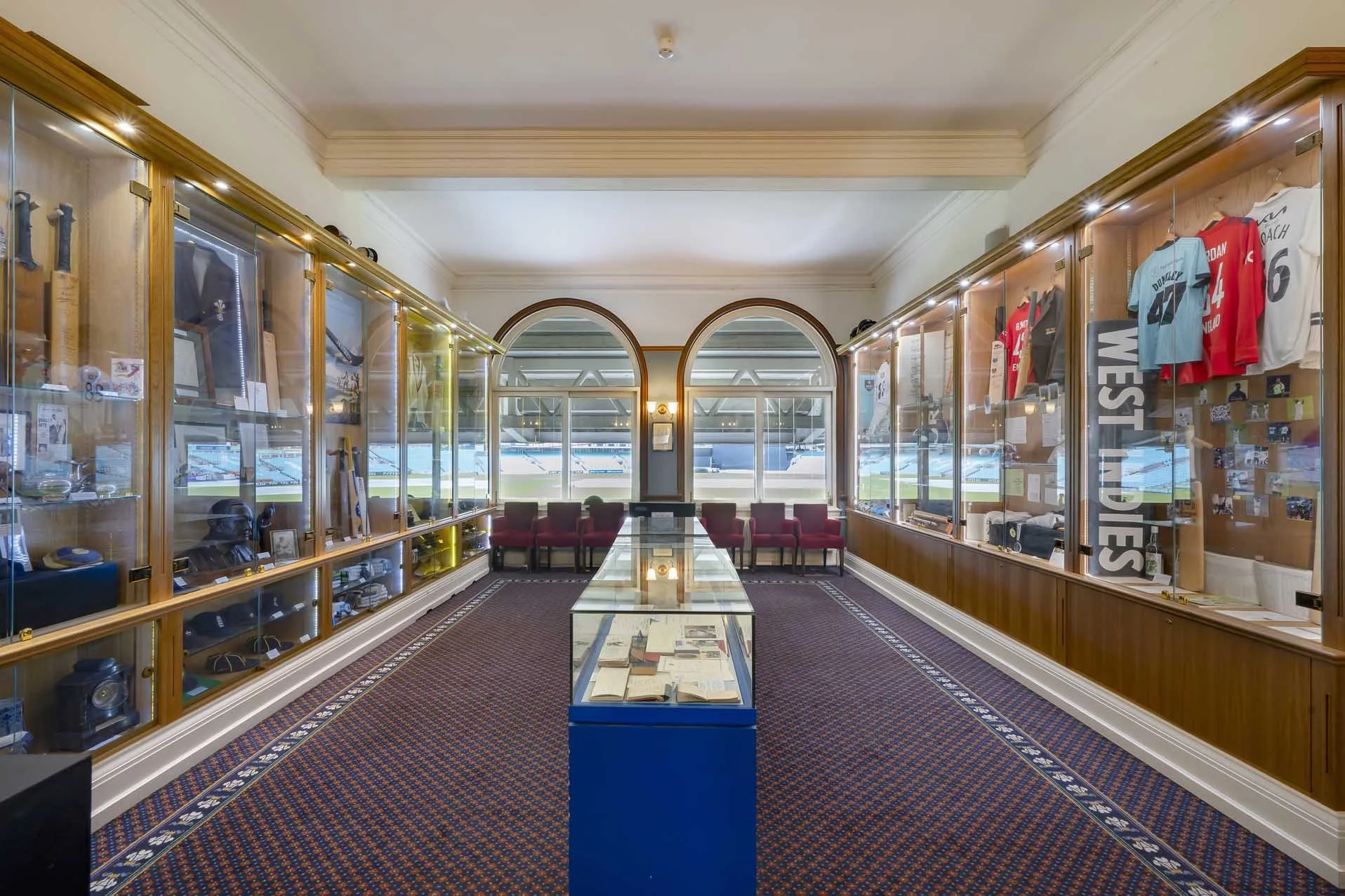 Room inside a sports stadium showcasing memorabilia and jerseys in glass display cases, with chairs along the window and a glass table in the center.