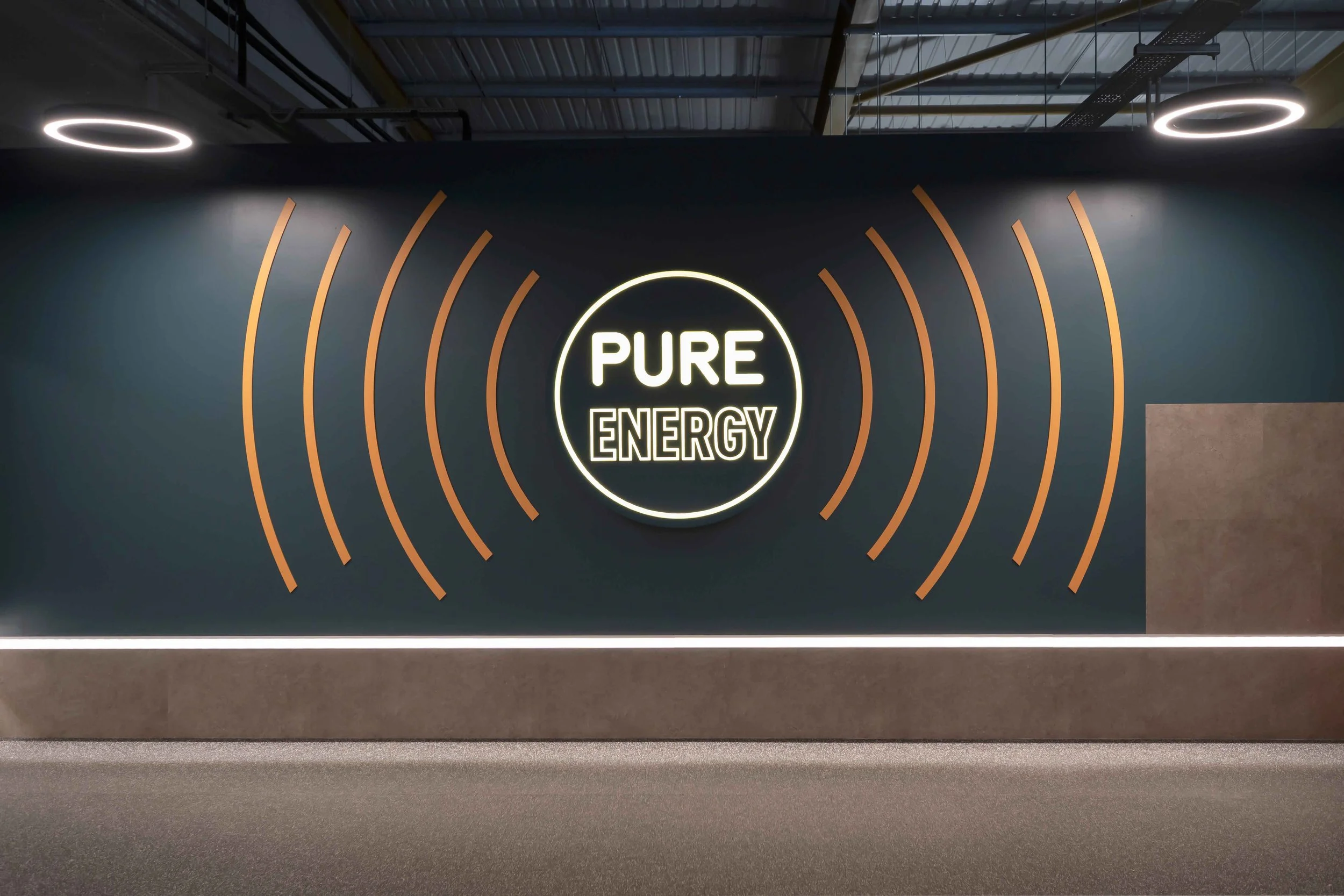 A wall with a neon sign that reads 'PURE ENERGY' surrounded by orange curved lines, with a ceiling featuring exposed pipes and circular lights.