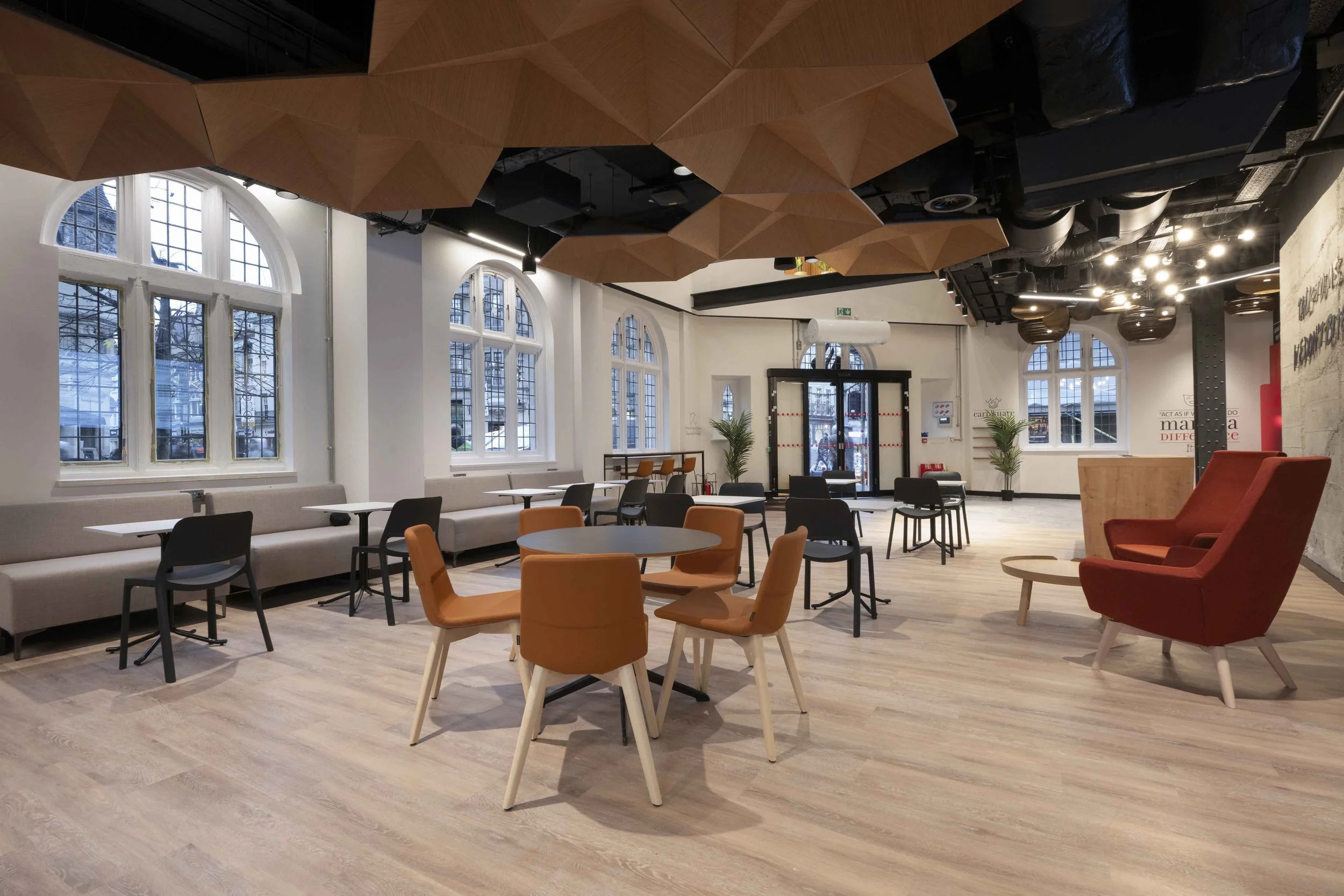 Spacious modern cafe with large arched windows, wooden ceiling panels, and a variety of colorful chairs and tables, illuminated by ceiling lights.