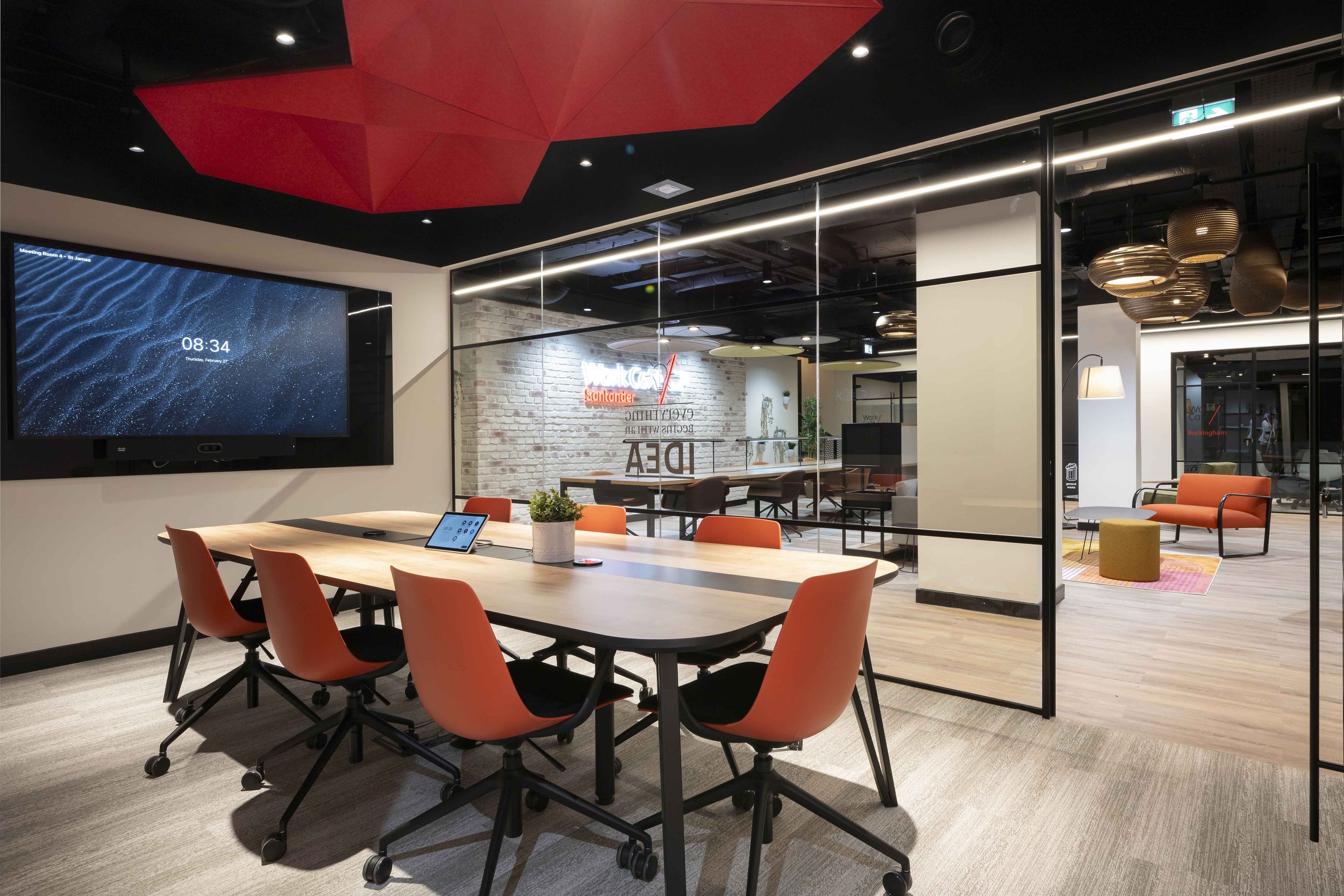 Modern conference room with a large table surrounded by orange chairs, a wall-mounted screen displaying the time, and a glass wall revealing a lounge area with stylish lighting, seating, and a brick wall with neon signage.